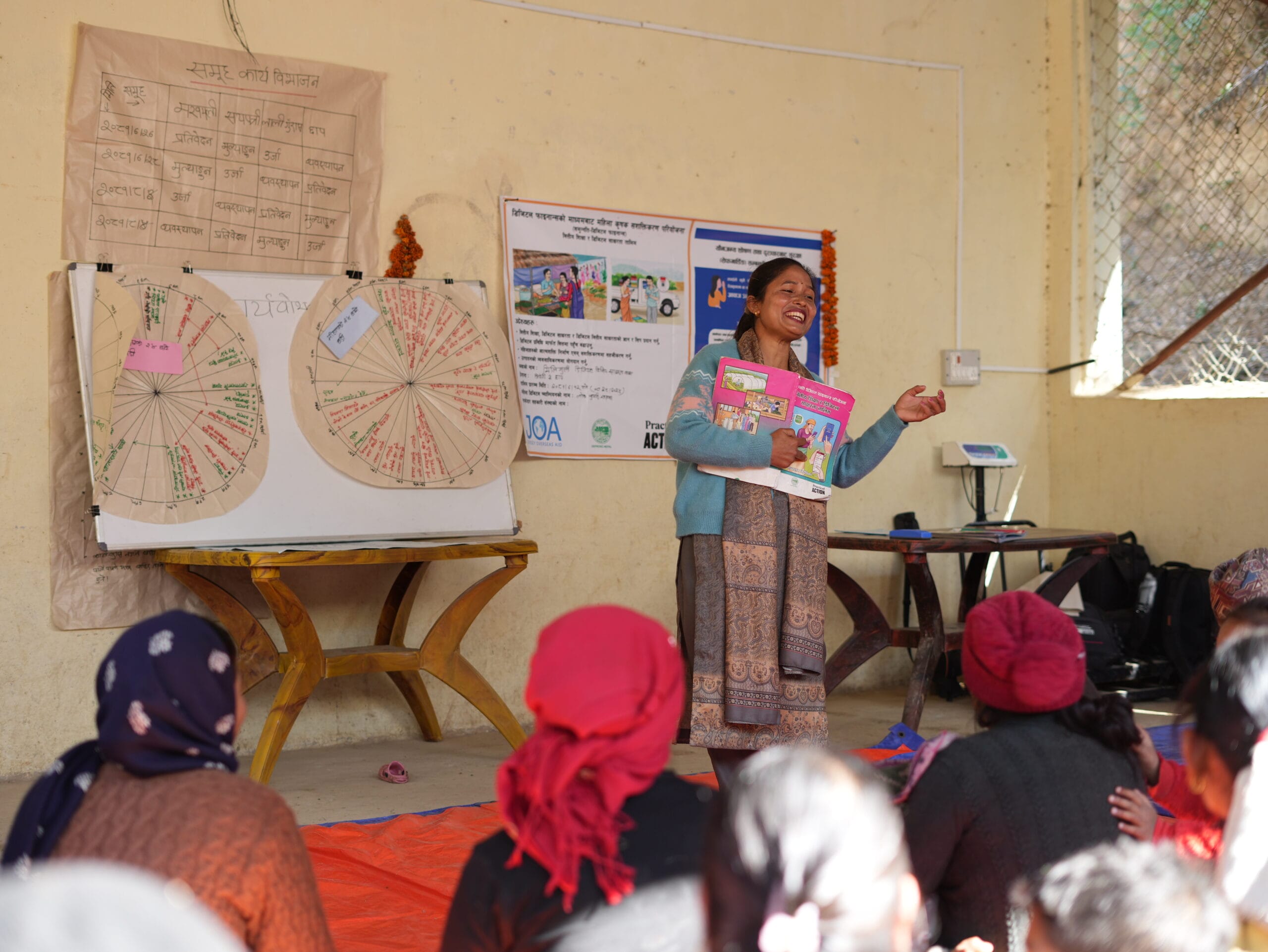 A woman stands and speaks in front of a small group, holding colourful booklets. Behind her are charts and posters with writing and diagrams in Hindi, displayed on the wall. The group sits on mats, listening attentively.