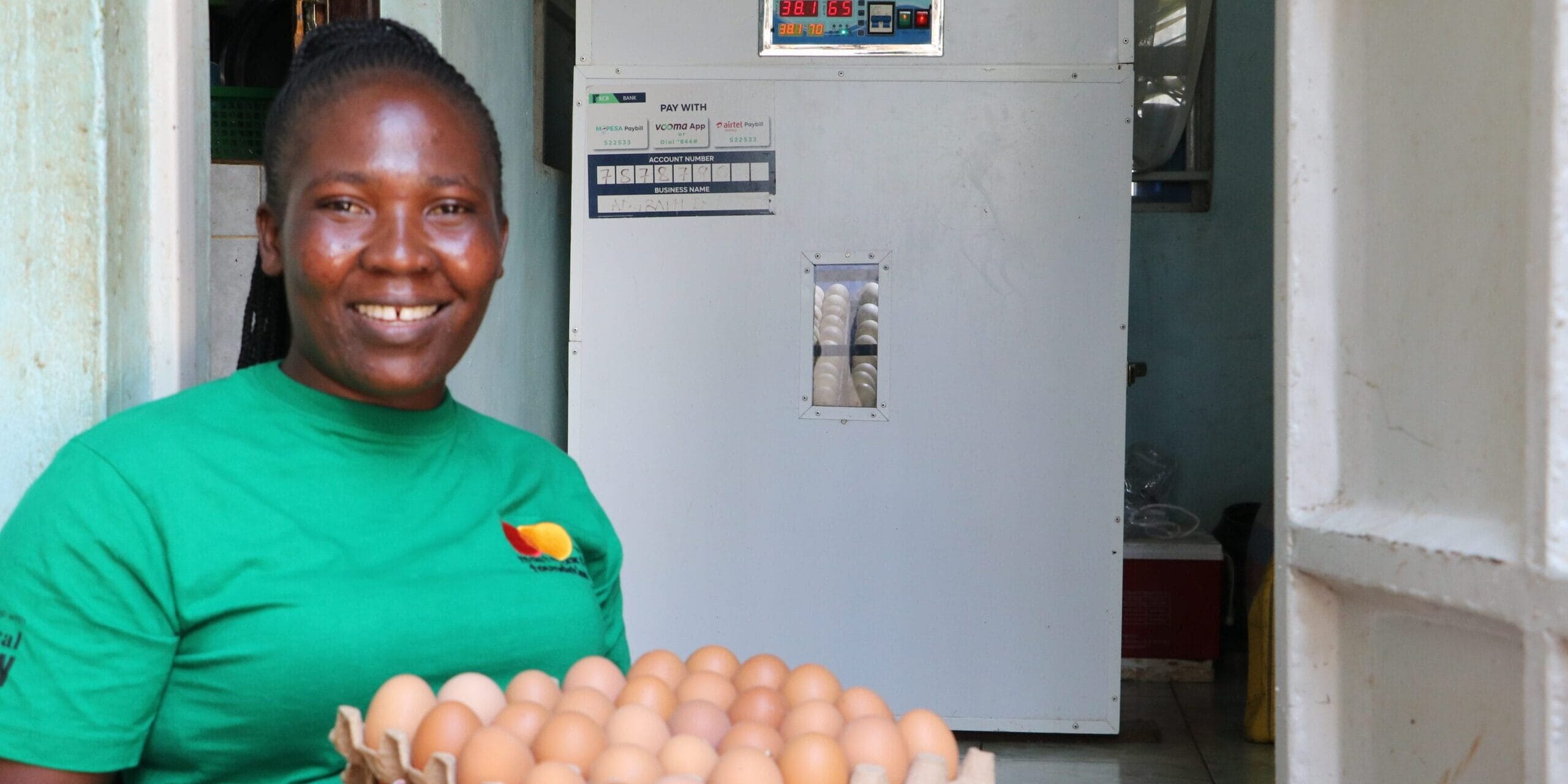 A woman in a green shirt smiles whilst holding a tray of eggs. Behind her is an egg incubator inside a small room.