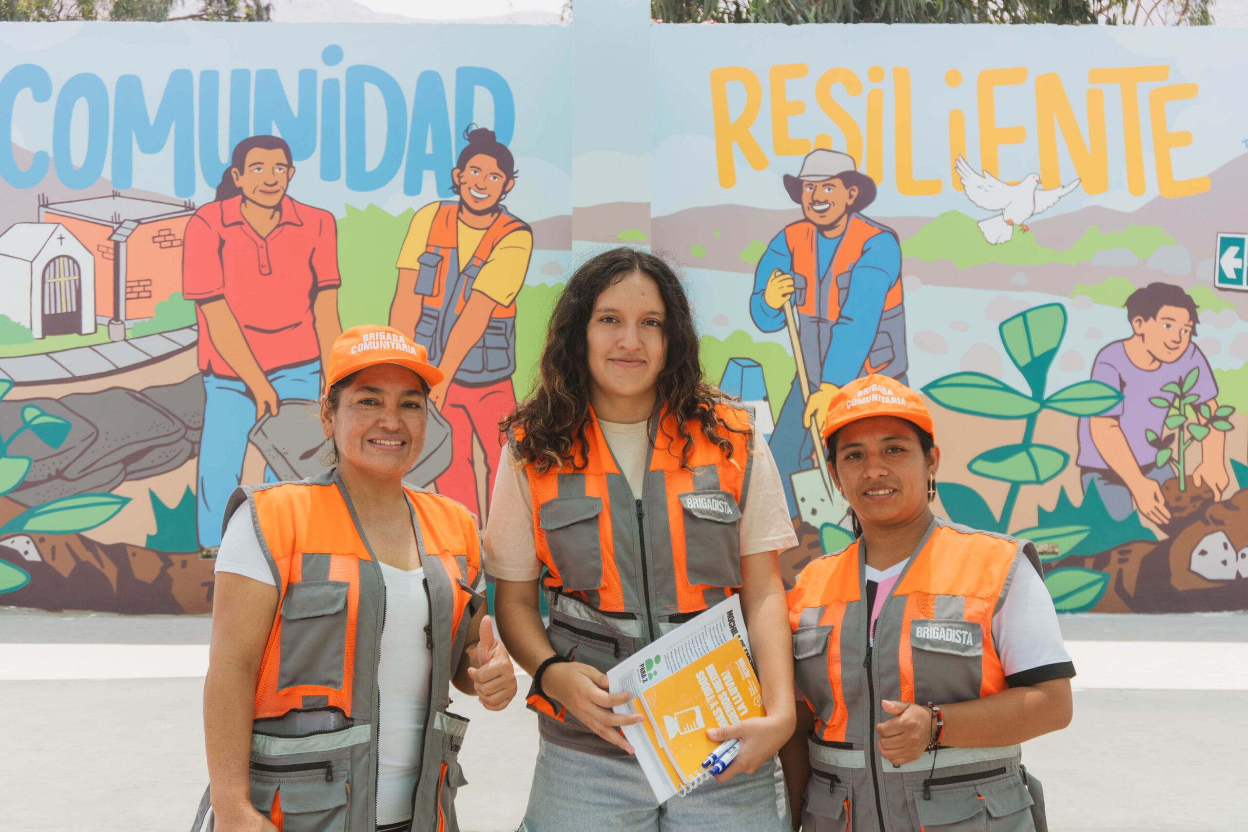 Three women wearing orange safety vests and hats smile and pose for a photo. A colourful mural behind them reads “COMUNIDAD RESILIENTE” and depicts people working together in a community garden.