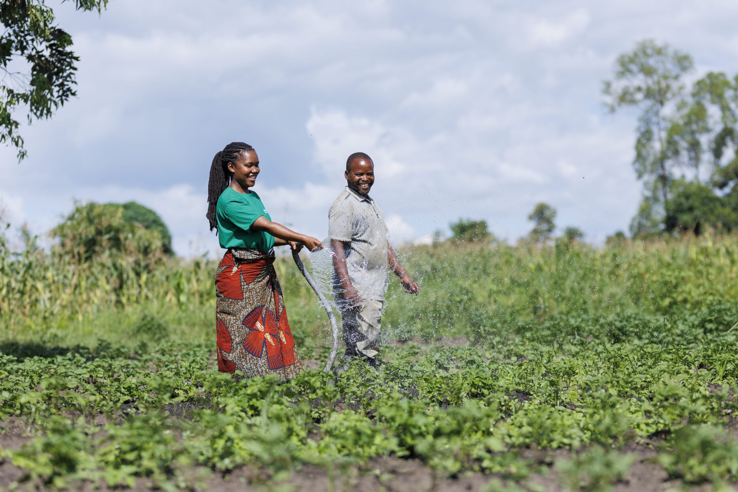 A woman and a man stand in a green field, smiling and watering crops with a hose under a bright sky. Tall plants and trees are visible in the background.