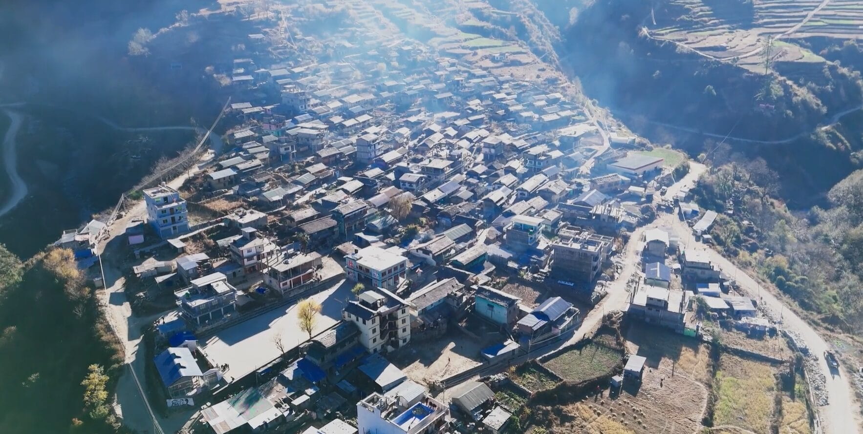 Aerial view of a densely packed village with rectangular buildings surrounded by mountains and terraced fields, bathed in sunlight with a visible haze or smoke above the rooftops.
