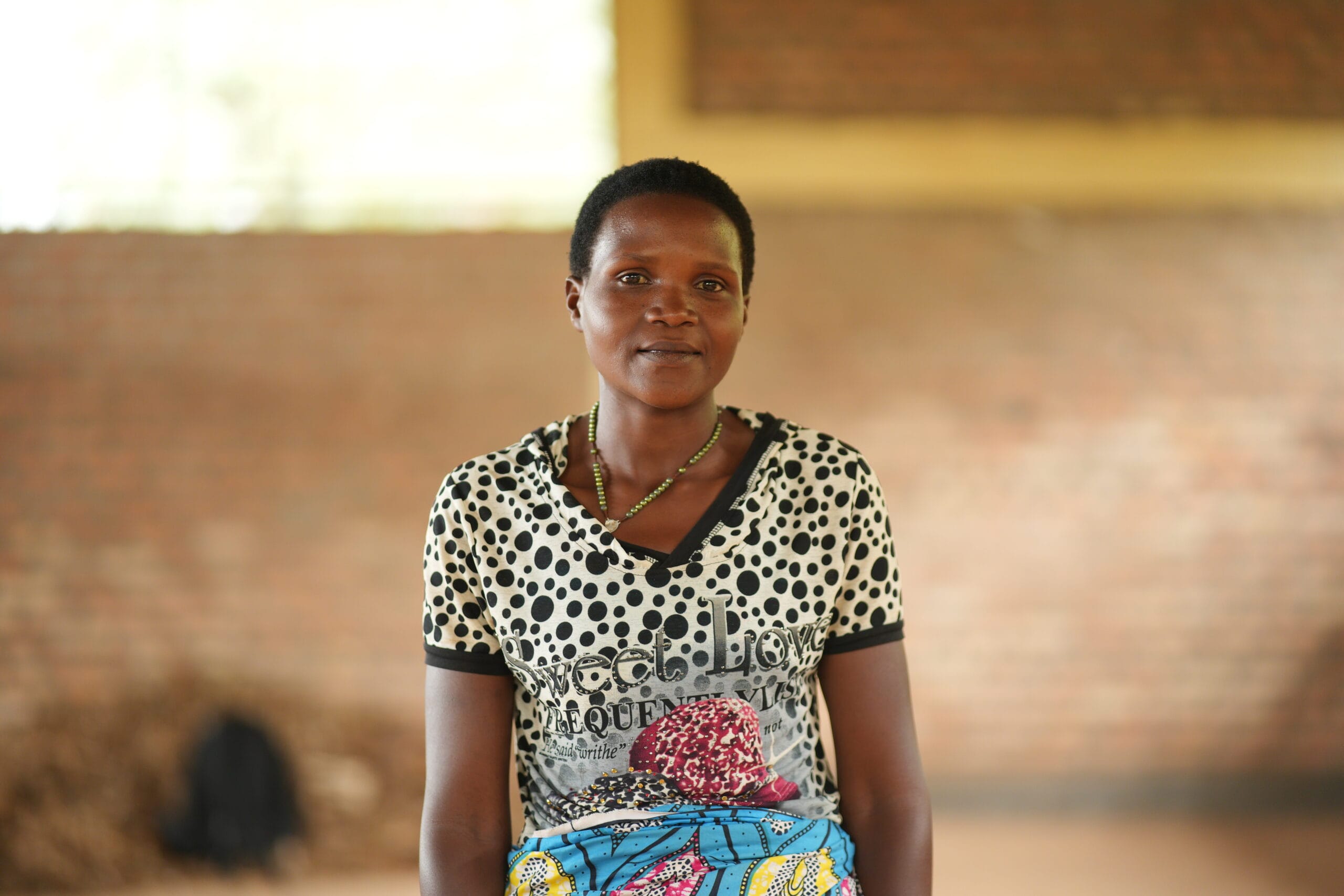 A woman with short hair wearing a black and white polka-dot shirt and a colourful skirt stands indoors in front of a blurred brick wall, looking calmly at the camera.