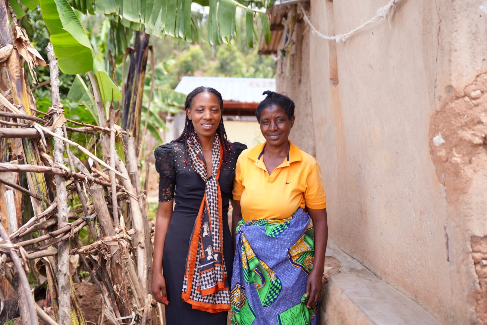 Two women stand side by side outdoors, one wearing a black dress with a colourful scarf, the other in an orange shirt and patterned wrap skirt. They are smiling, with a mud wall and banana plants in the background.