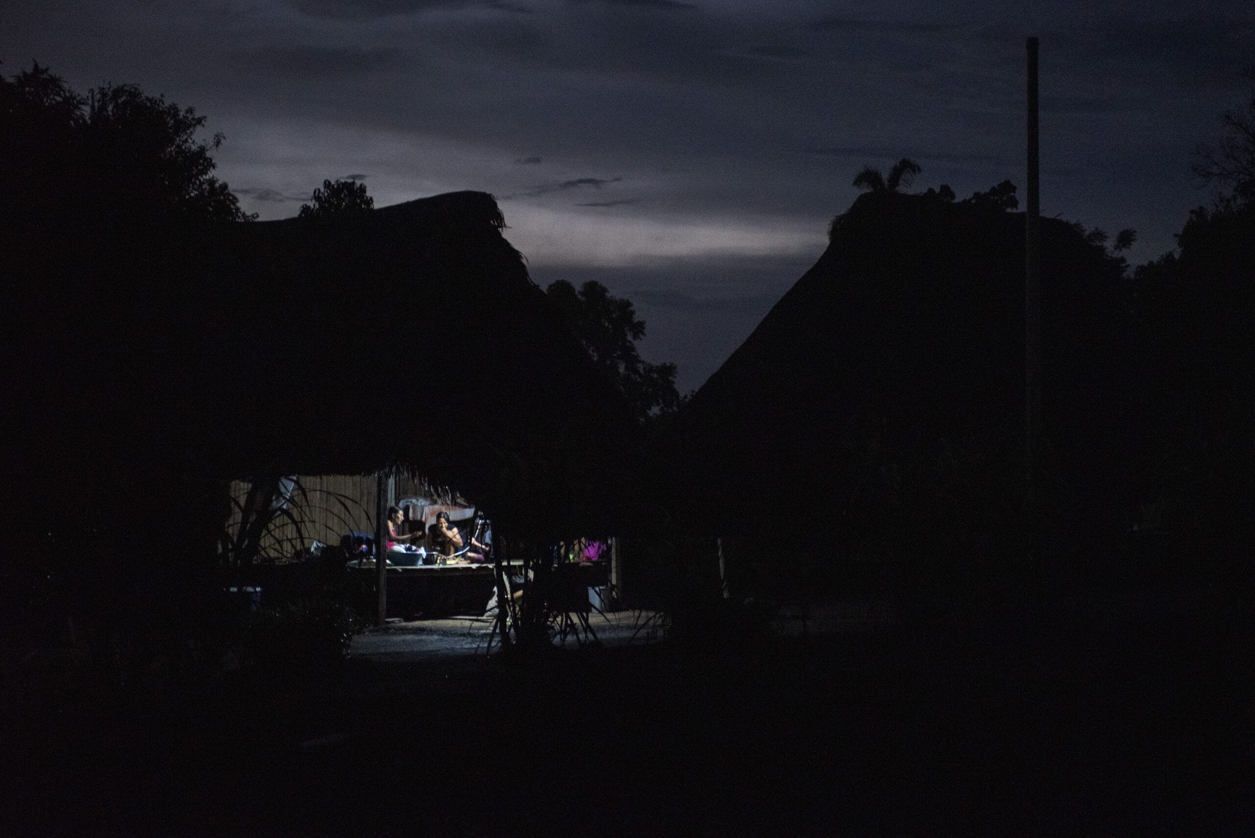 A dimly lit thatched hut at night, with people gathered inside around a table. The surrounding area is dark, with silhouettes of trees and another hut visible against the evening sky.