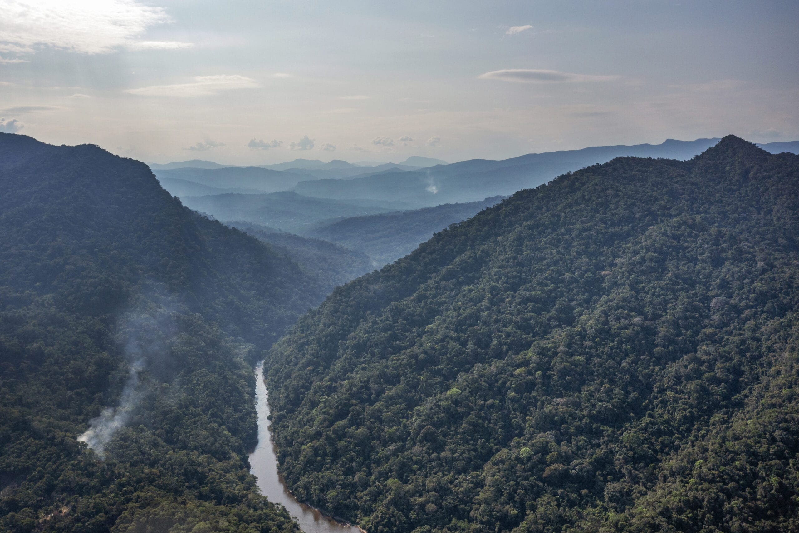 Aerial view of a dense, green rainforest with a winding river cutting through steep, forested hills under a cloudy sky, with distant mountains in the background and wisps of smoke rising from the trees.