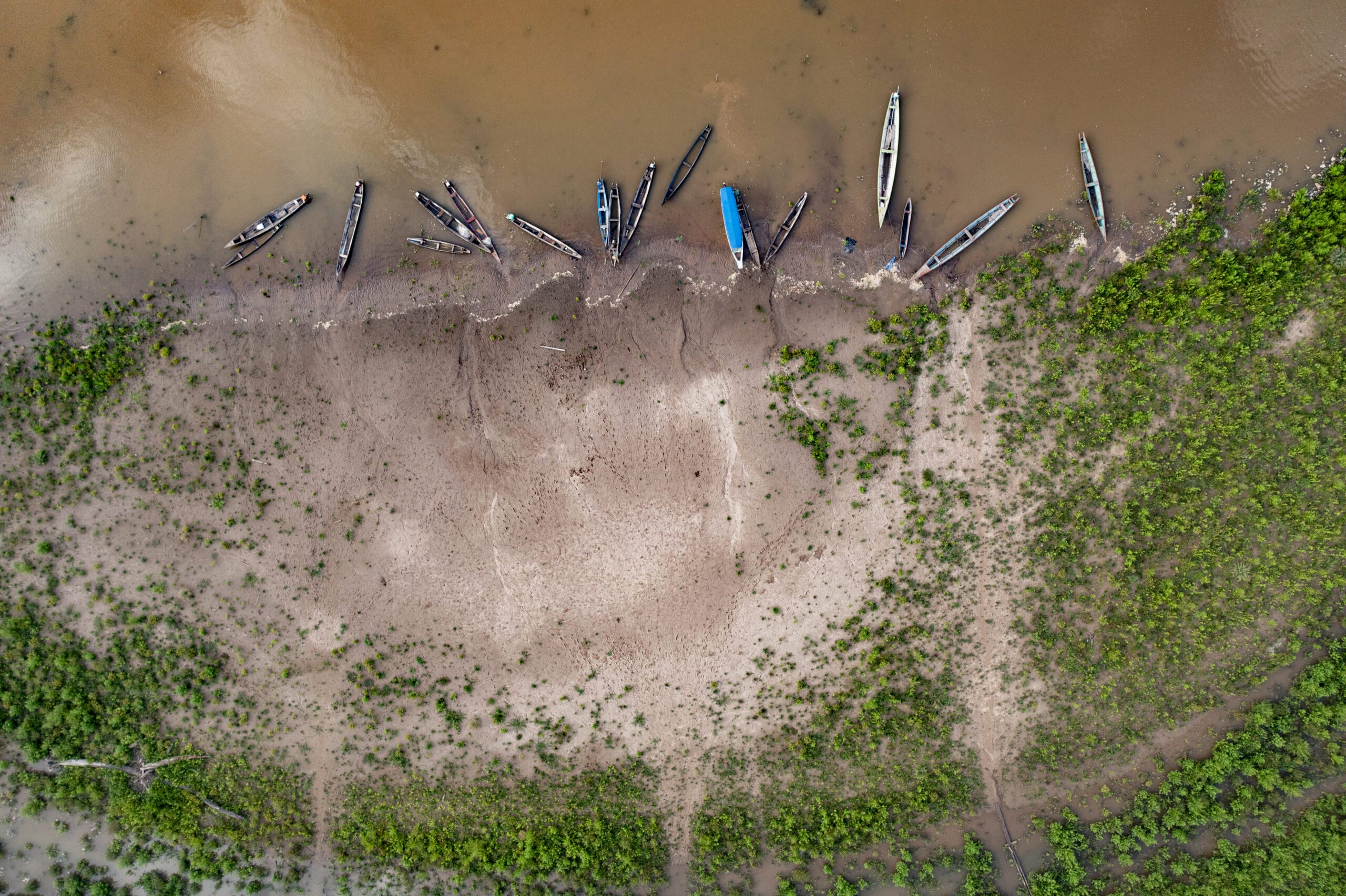 Aerial view of a muddy riverside with several wooden boats moored along the shore. Green vegetation borders the sandy, partially eroded riverbank, contrasting with the brown water.