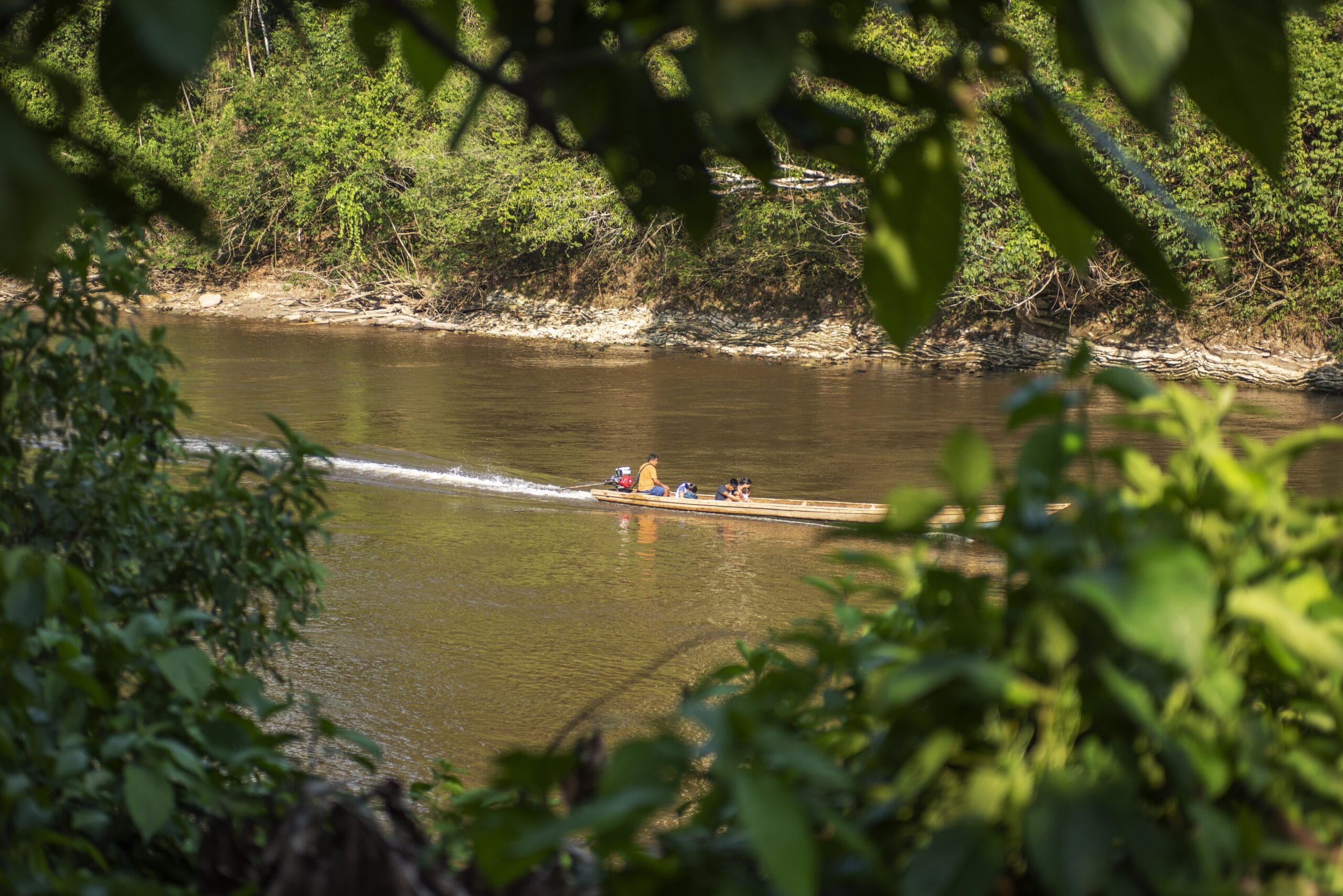 A small wooden boat with several people travels down a calm river, surrounded by lush green forest, as seen through an opening in dense foliage.