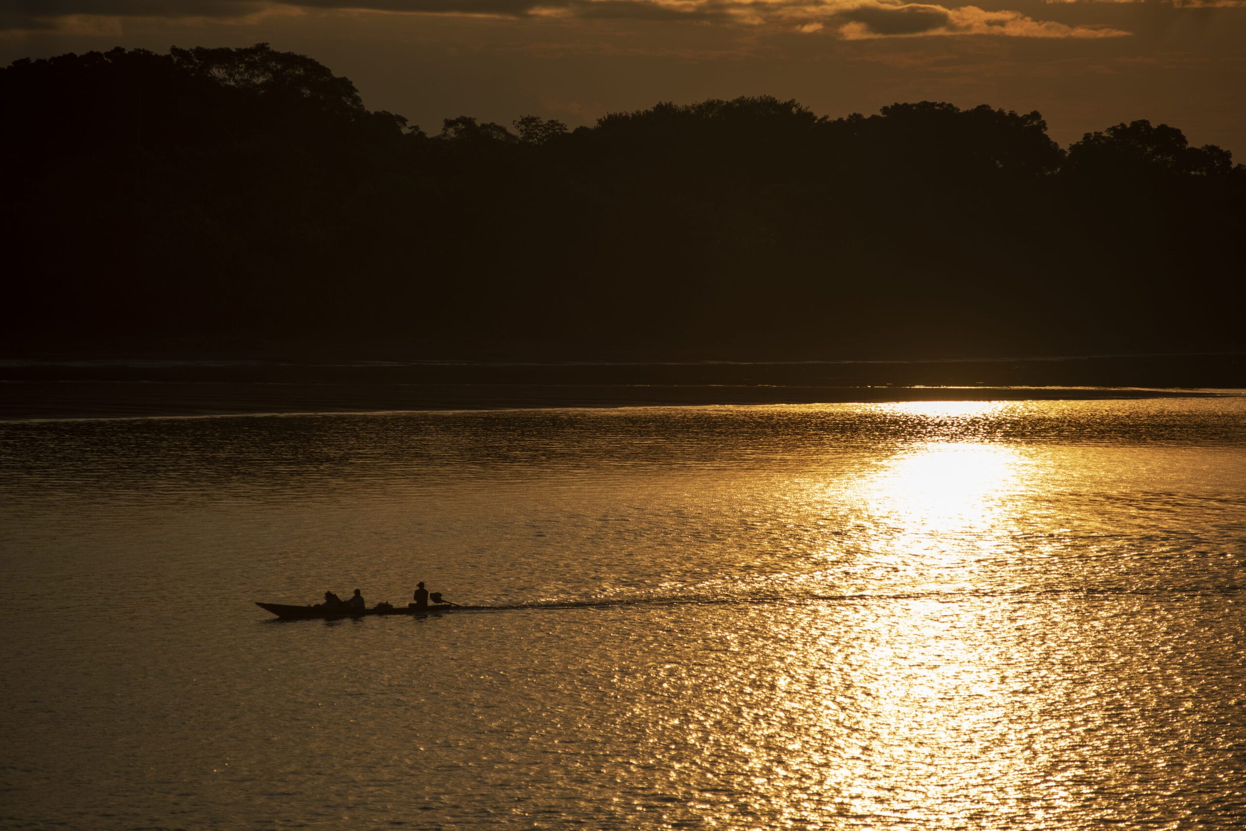 A small boat with three people glides across a calm river at sunset, casting ripples over the golden, shimmering water with dark silhouettes of trees in the background.