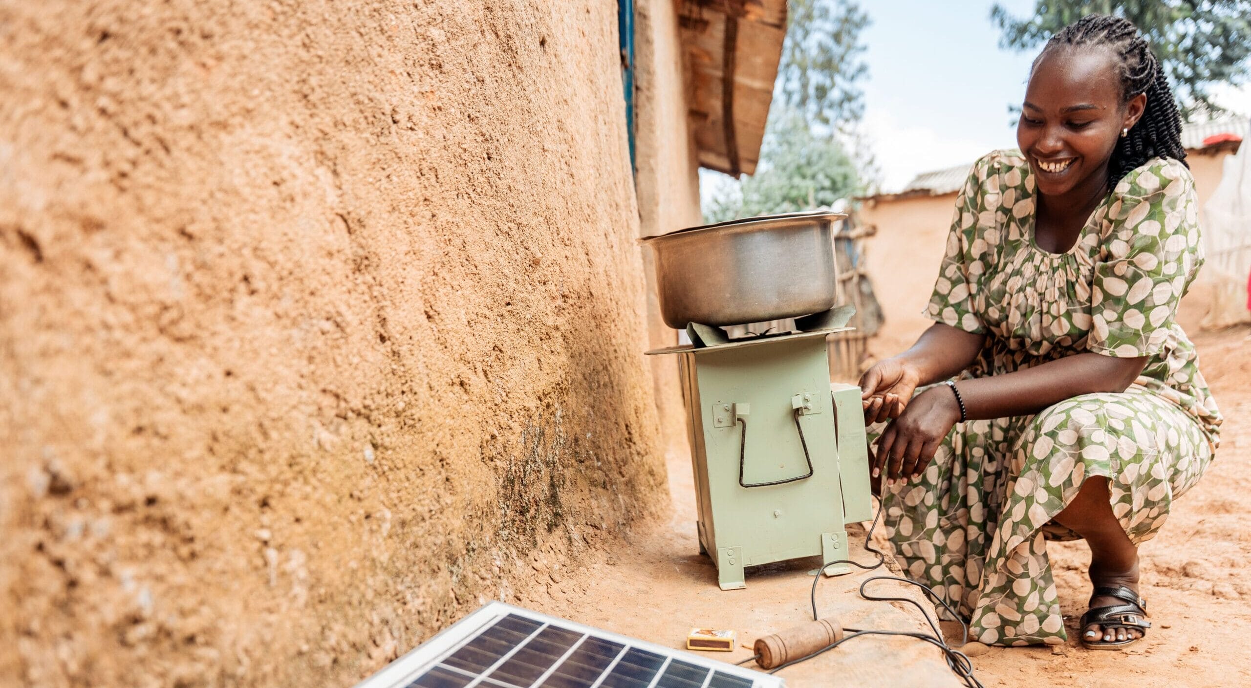 A woman in a patterned dress smiles as she squats beside a solar panel connected to a green cooking stove outdoors, near an earthen wall in a rural setting.