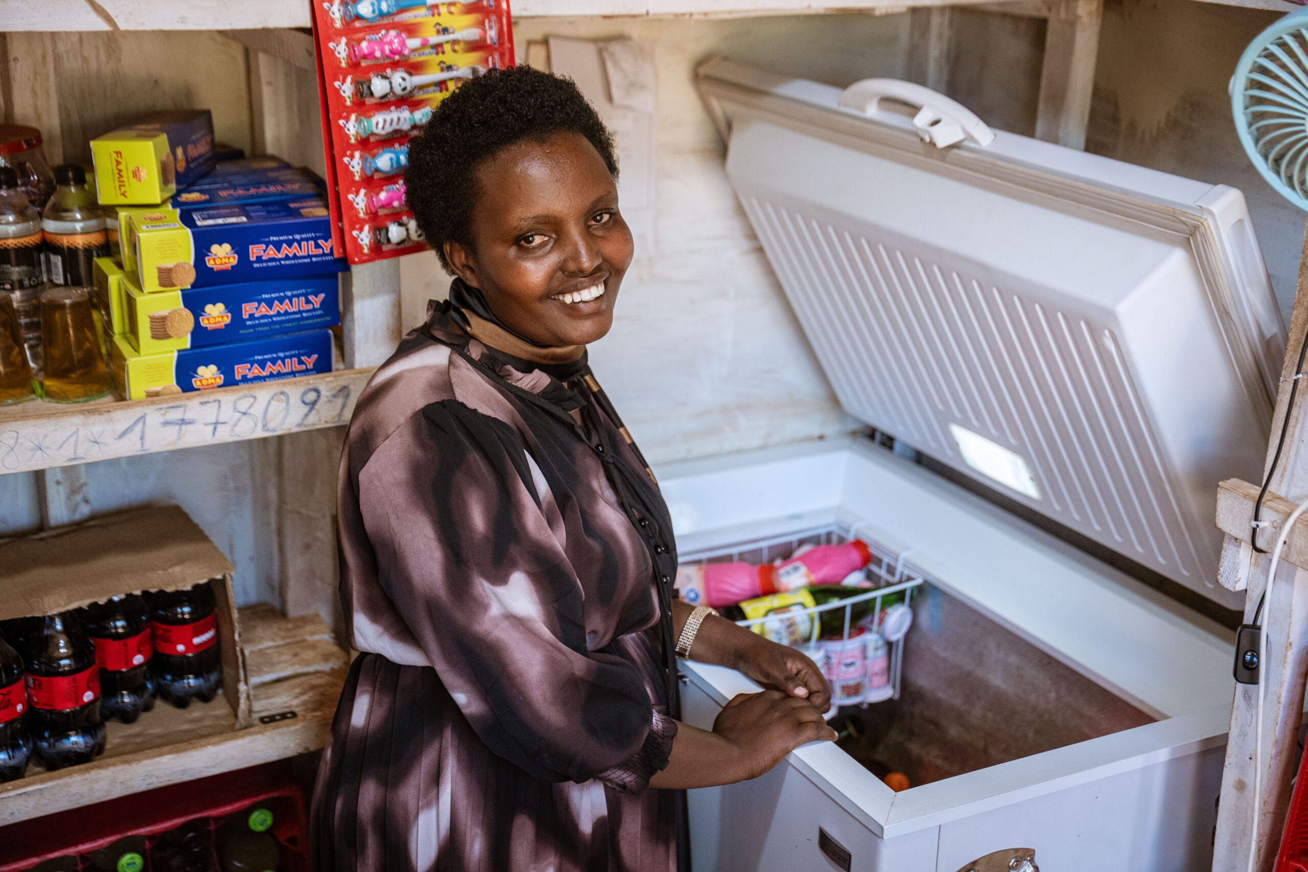 A woman smiles while standing next to an open chest freezer in a small shop, surrounded by shelves stocked with drinks, cooking oil, and snack items.