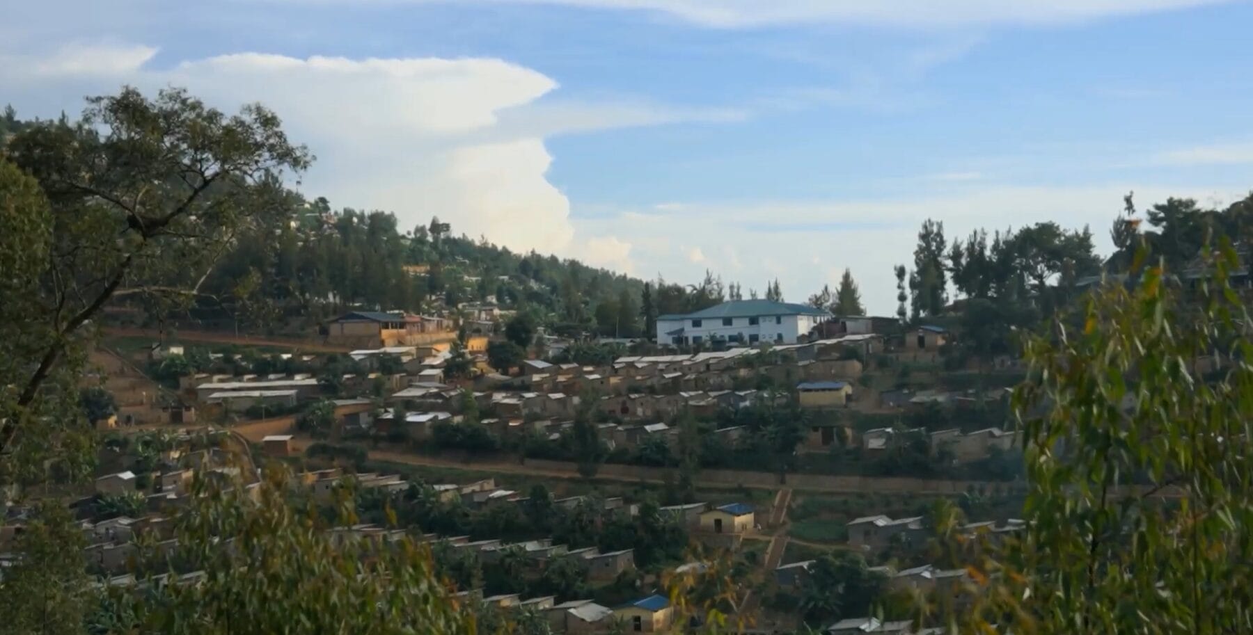 A hillside settlement with many small houses and a large white building surrounded by trees, under a partly cloudy sky.