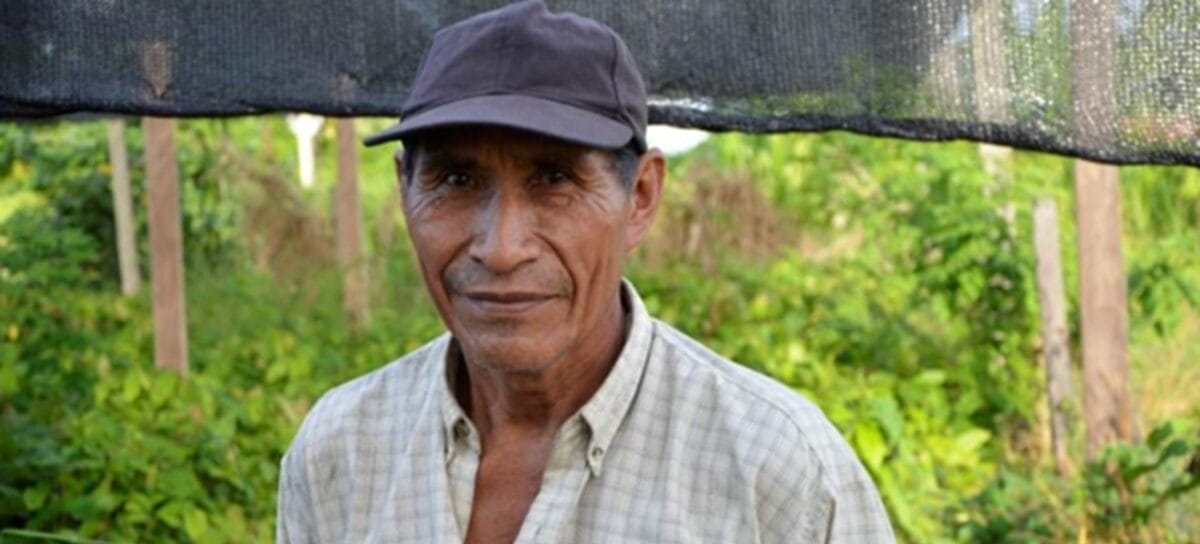 An older man wearing a dark cap and a light checked shirt stands outdoors in front of lush green vegetation, looking at the camera with a neutral expression.