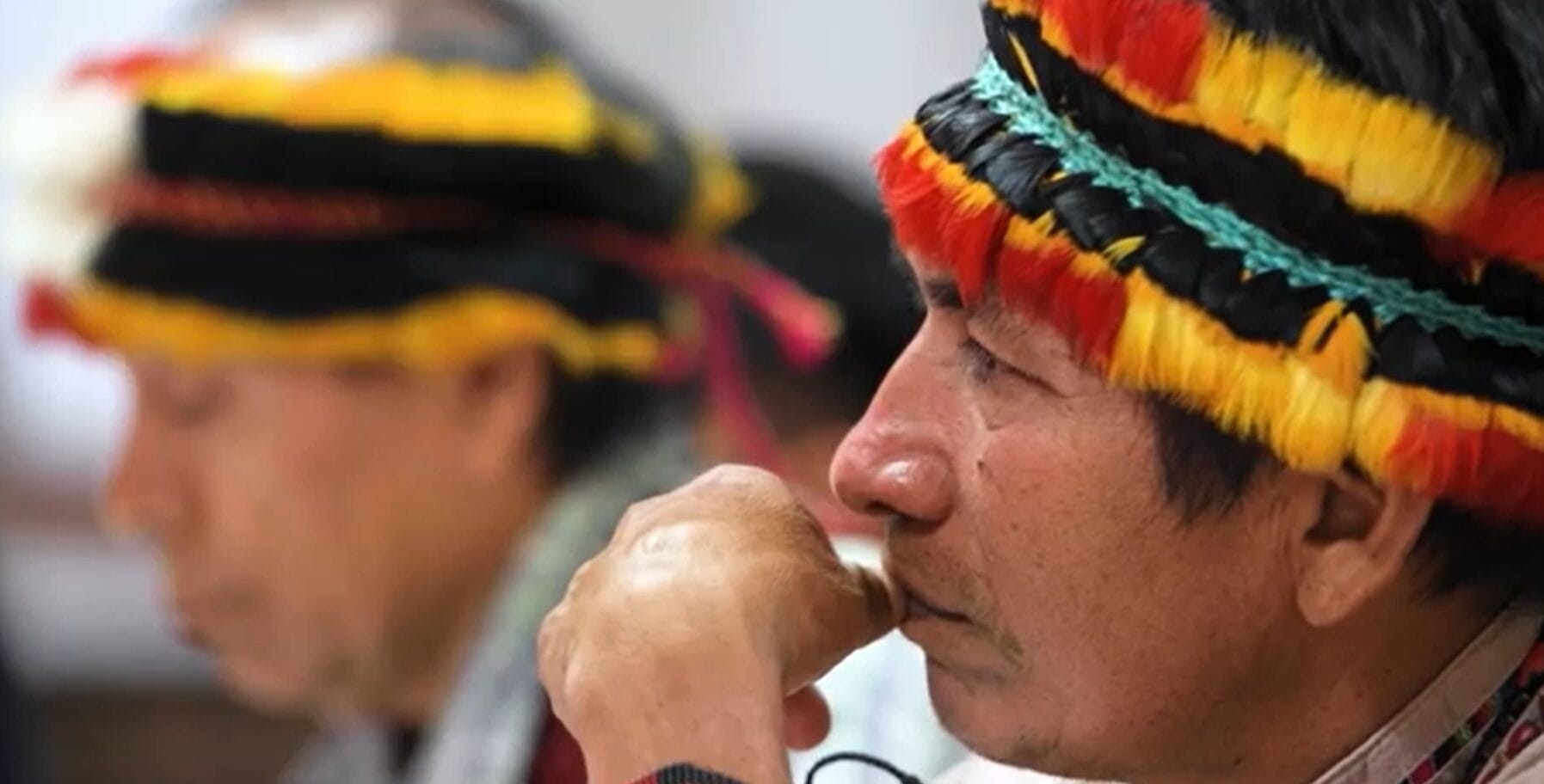 Two men in profile wear traditional feathered headbands in black, red, and yellow. The man in the foreground rests his chin on his hand, whilst the man in the background is out of focus.