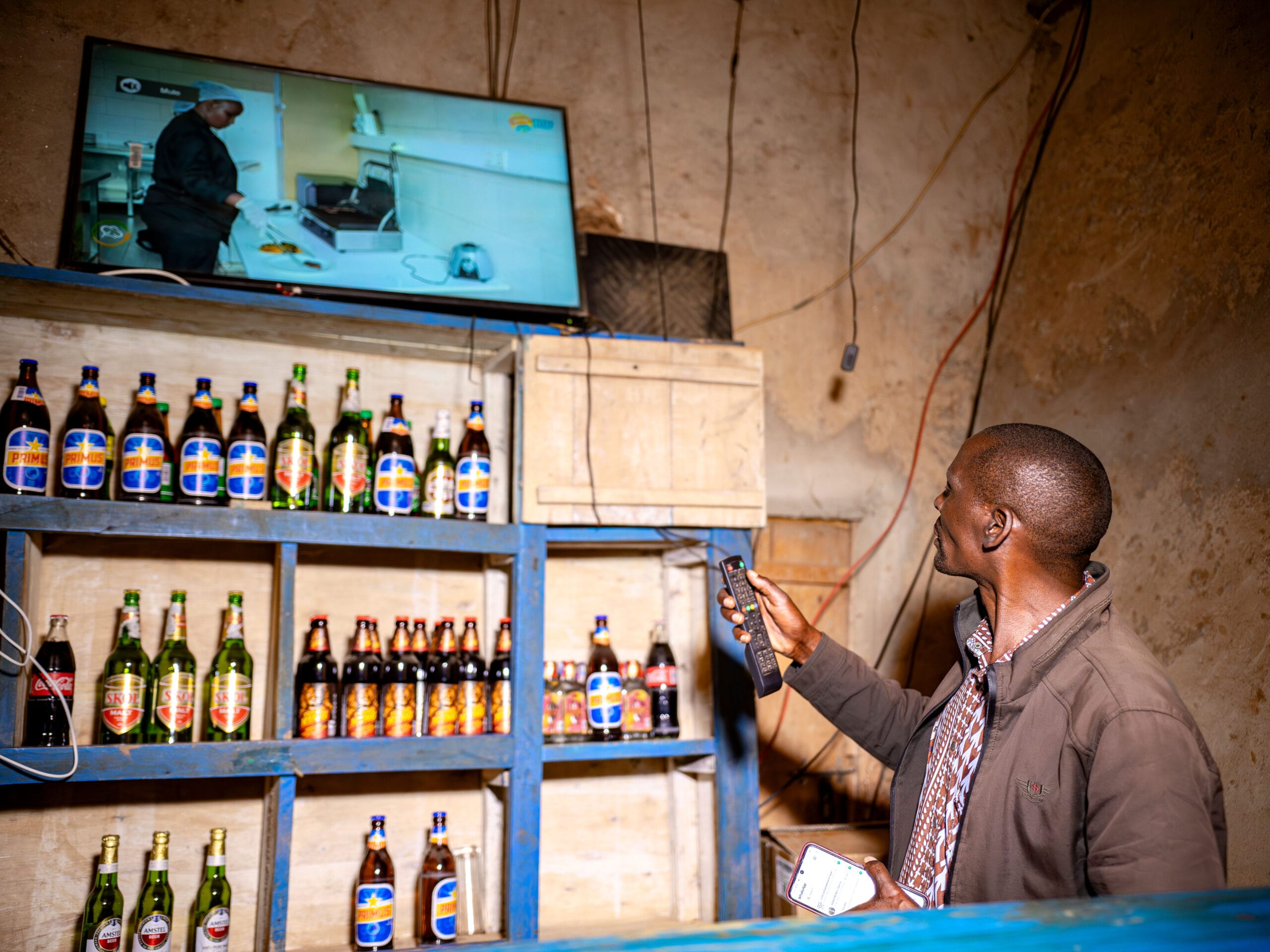 A man stands behind a bar shelf filled with assorted bottles, holding a remote and a mobile, whilst watching a telly mounted on the wall showing a cookery programme.