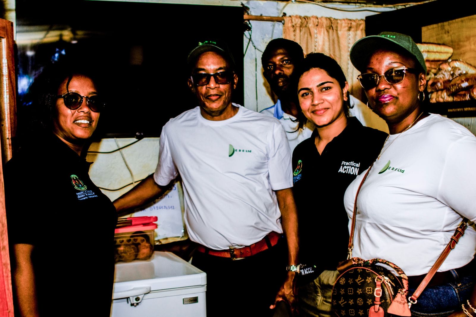 Five people stand indoors, smiling at the camera. Four wear caps and casual clothing, two in white T-shirts, two in black, with “Practical Action” visible. A freezer, files, and a TV are seen in the background.