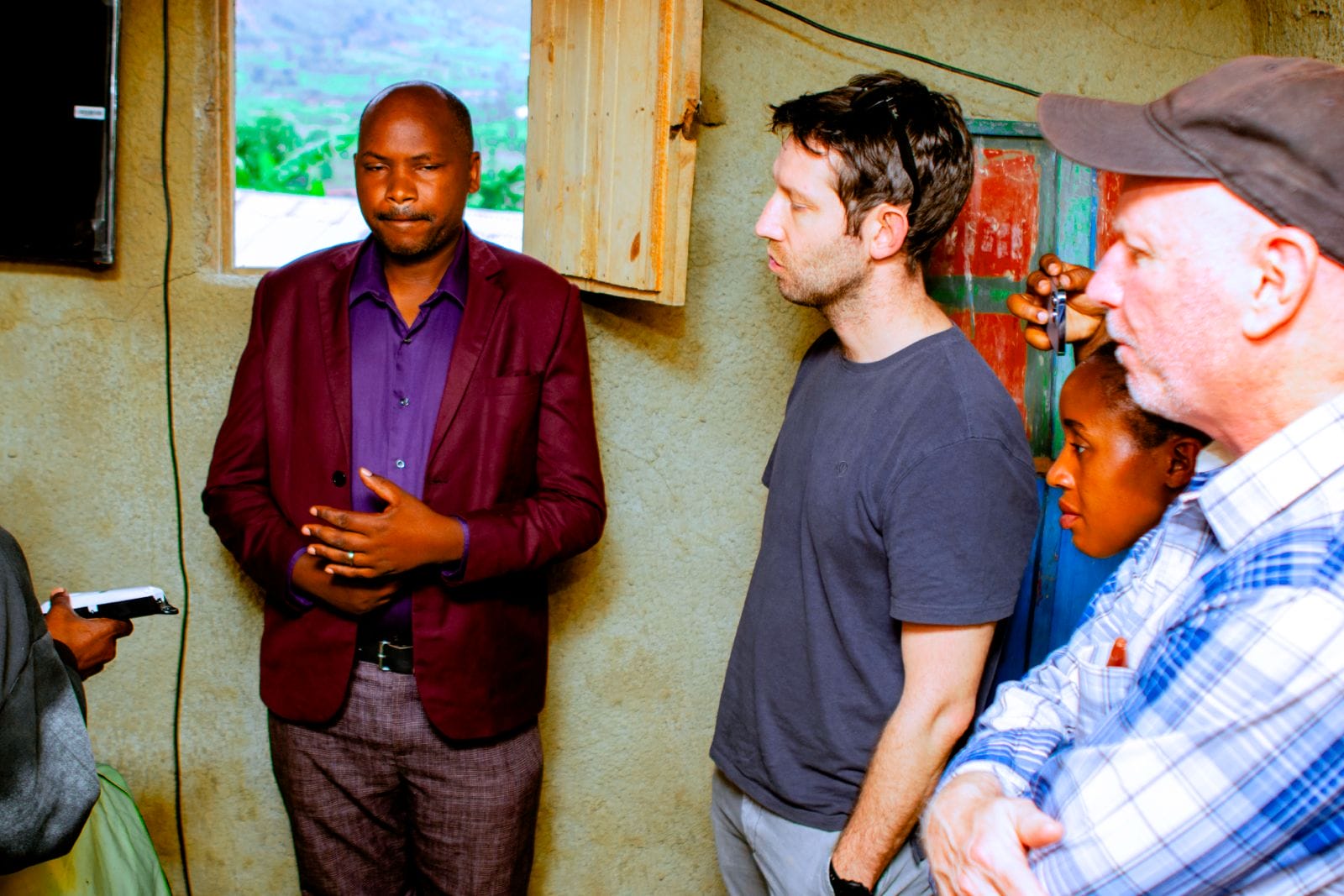 Four people stand indoors having a discussion. One man in a purple jacket speaks whilst three others, including a woman and two men, listen attentively. A window and a partially open door are visible in the background.