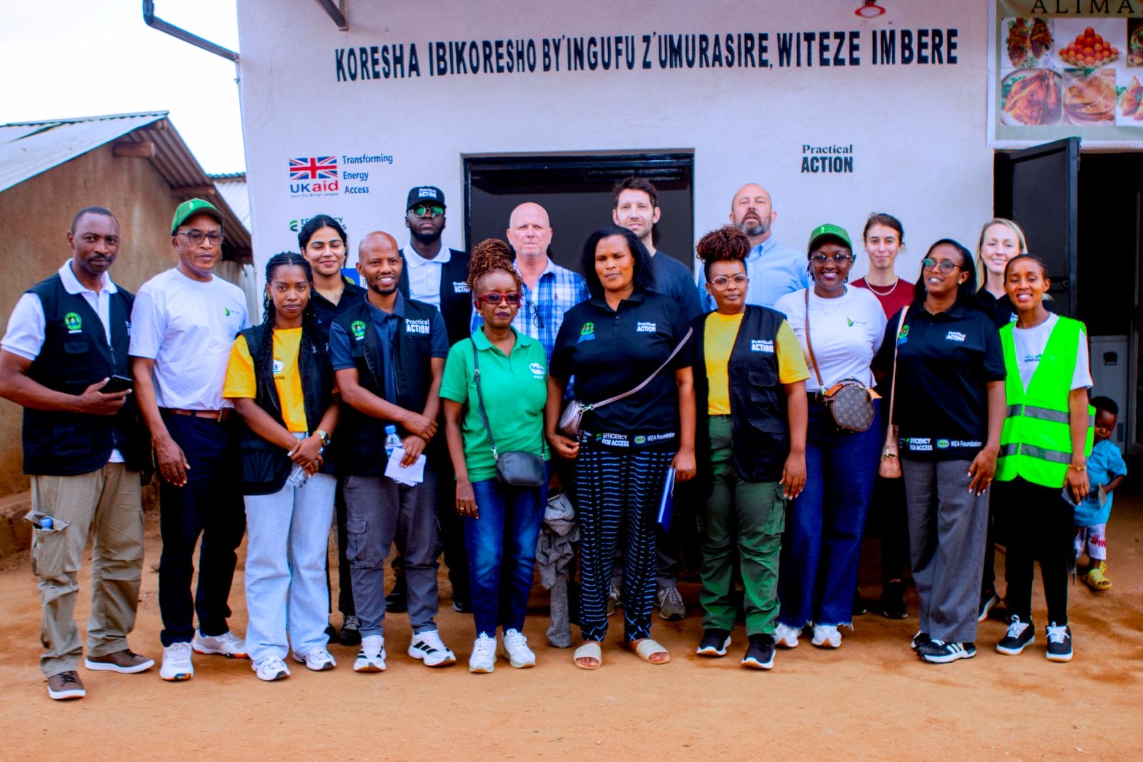 A diverse group of people stands and poses for a photo in front of a building with a sign in Kinyarwanda and English, some wearing black waistcoats and green uniforms, smiling at the camera.