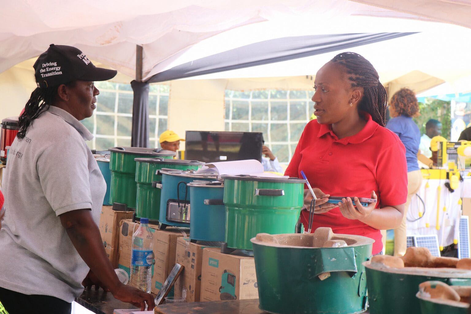 Two women talk at a stand displaying green and blue cooking stoves. One woman wears a red shirt and holds a notebook, while the other wears a grey shirt and black hat. Other people and equipment are visible in the background.