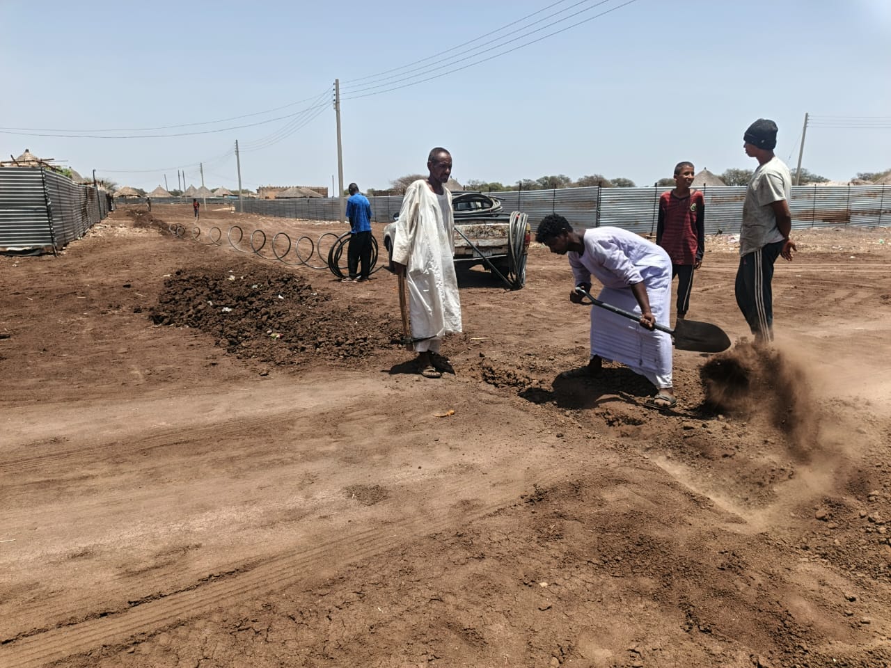 Several people work together on a dusty dirt road, shovelling soil. Metal fencing and utility poles line the sides, with corrugated metal sheets and pipes visible in the background under a clear sky.