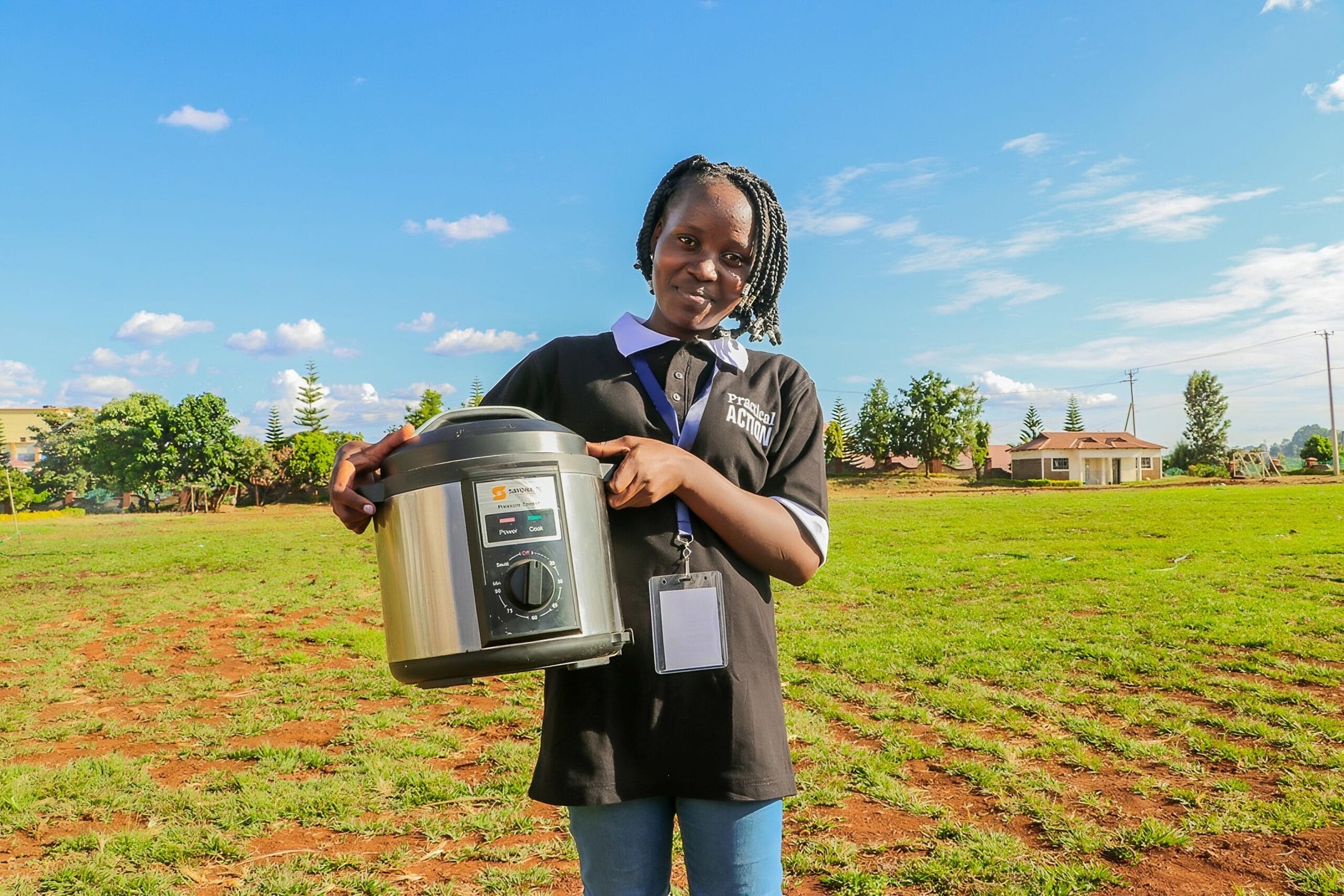 A young woman stands on a grassy field holding an electric pressure cooker, smiling at the camera. She wears a black shirt, a lanyard with a badge, and there are buildings and trees in the background under a blue sky.
