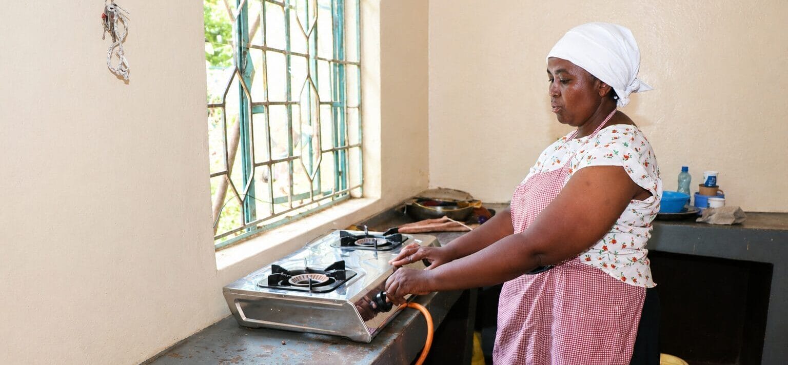 A woman wearing a headscarf and apron stands in a kitchen, adjusting a portable gas cooker on a metal worktop near a window with bars. Cooking utensils and gas cylinders are visible in the background.