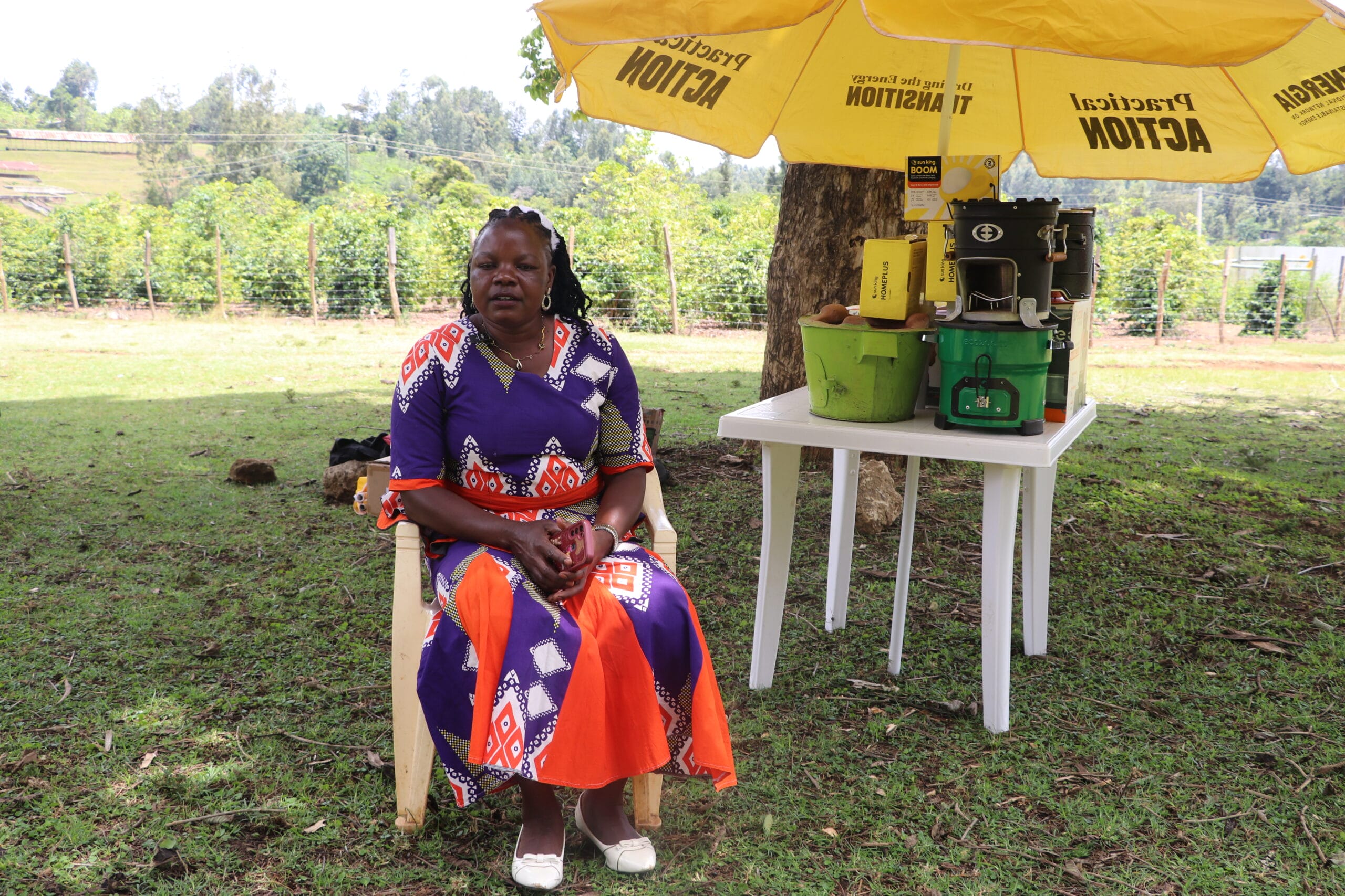 A woman in a colourful dress sits on a plastic chair outdoors under a yellow umbrella. Next to her is a white table with a green bucket, a juicer, and other items. Trees and greenery are visible in the background.