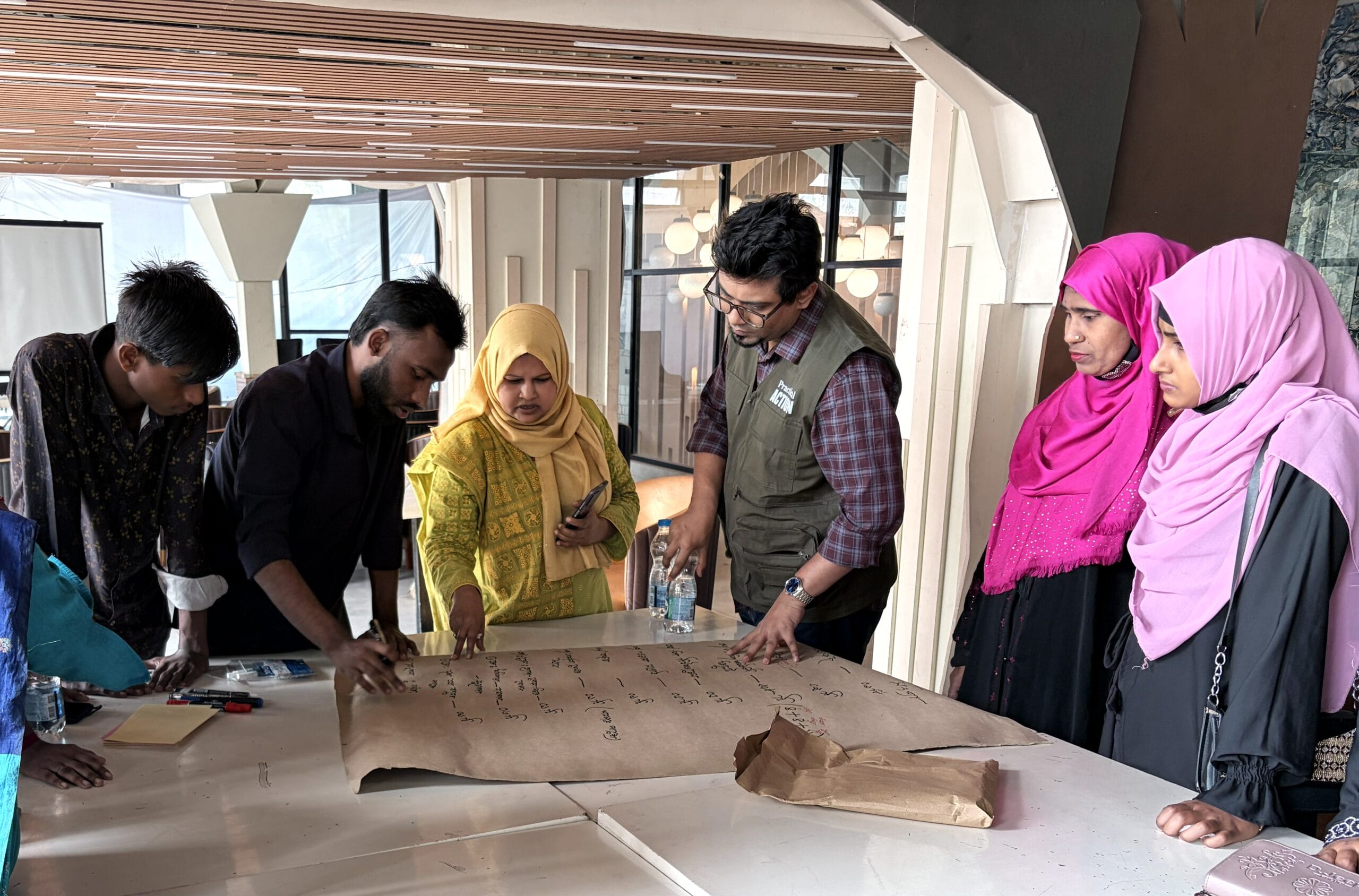 A group of people gathers around a table, closely examining and discussing notes written on a large sheet of paper. Some individuals wear brightly coloured hijabs and appear engaged in conversation.