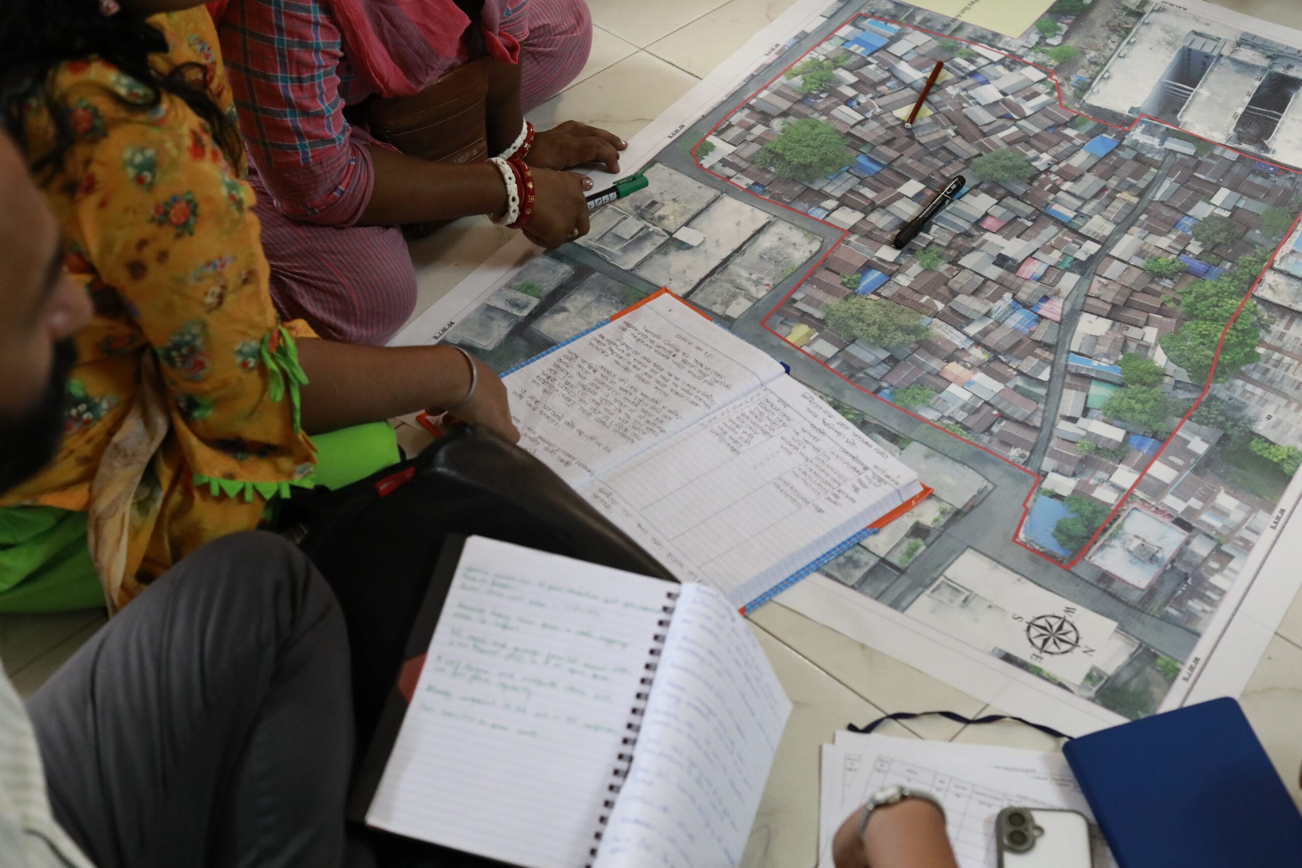 Several people sit on the floor around a large map of a densely built-up area, with notebooks and pens, discussing or taking notes, suggesting a planning or mapping activity.