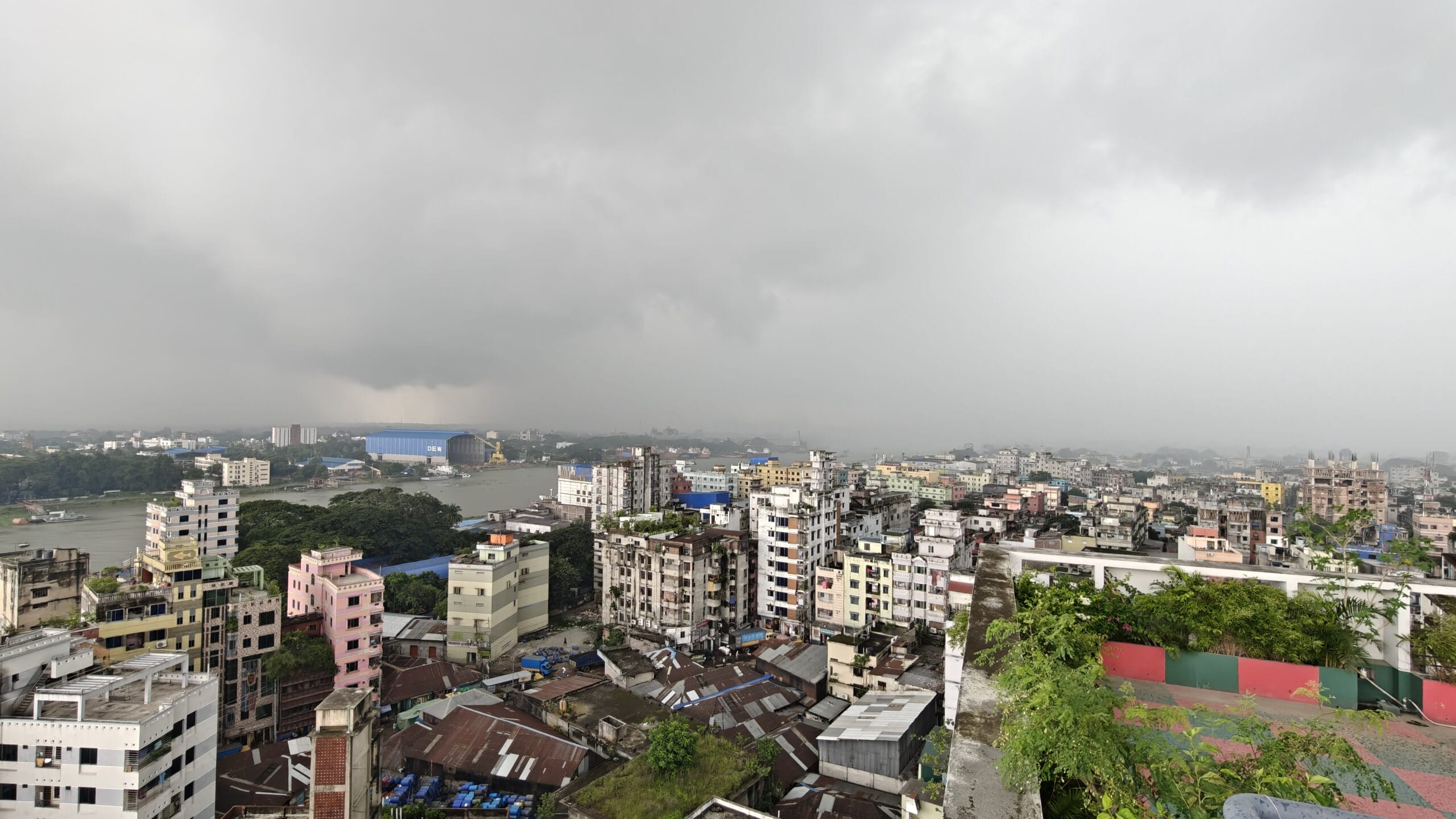 A cityscape under a cloudy sky, with densely packed buildings in various colours. A river flows through the city, and greenery is visible on some rooftops. The atmosphere looks misty or rainy.