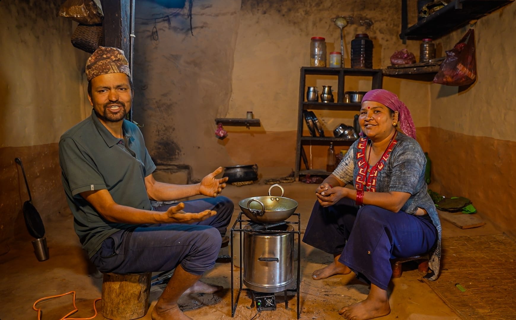 A man and woman sit on the floor of a rustic kitchen, smiling and talking beside a cooker with a cooking pot. The room has clay walls, shelves with kitchen items, and a warm, inviting atmosphere.