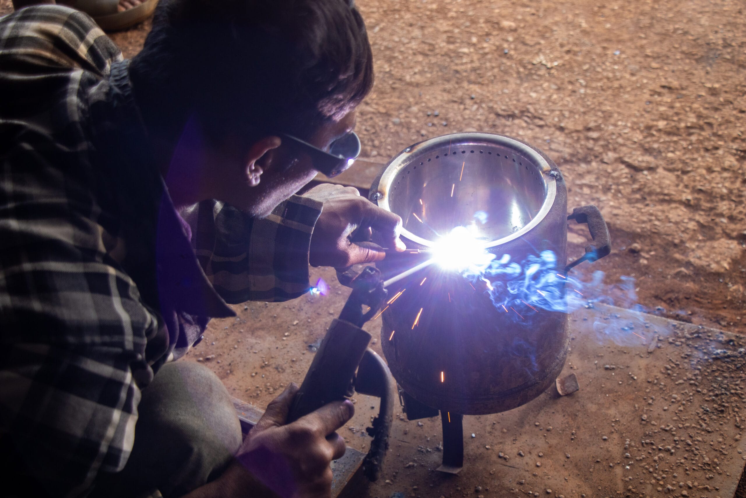 A person wearing protective glasses welds a metal object, creating bright sparks and smoke. The scene takes place on a rough, brown surface, and the person is dressed in a checked shirt.