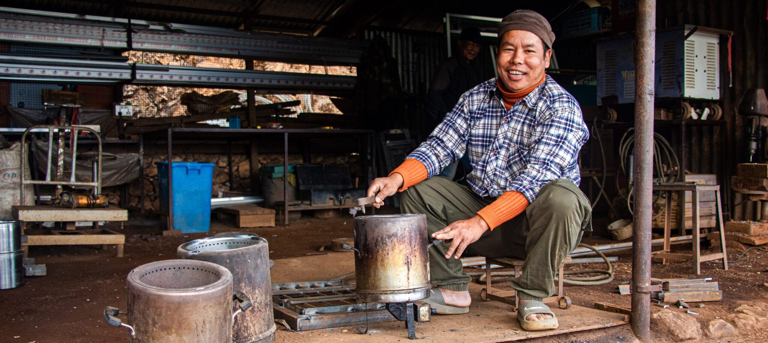 A smiling person wearing a checked shirt and hat is sitting on a stool in a workshop, holding a metal container over a cooker. Various metal equipment and tools are scattered around the rustic workspace.