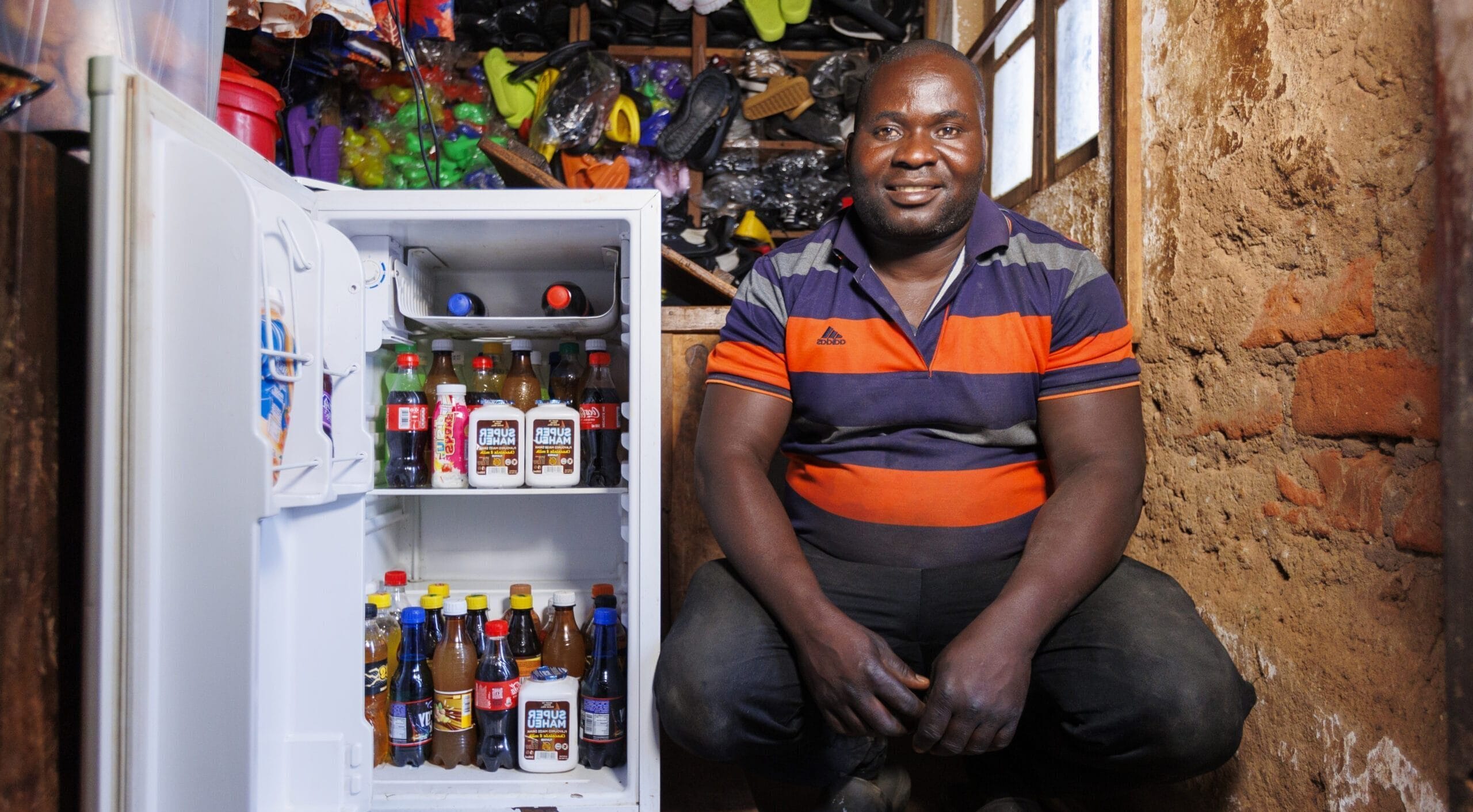 A man in a striped shirt squats next to an open fridge stocked with bottled drinks in a rustic room with brick walls. Shelves with colourful items are visible above and behind him.