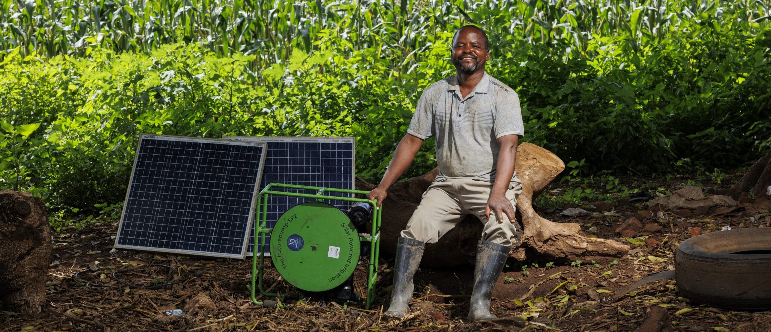 A man in wellington boots sits outdoors on a tree stump next to two solar panels and a green mechanical device, with lush greenery and tall maize plants in the background.
