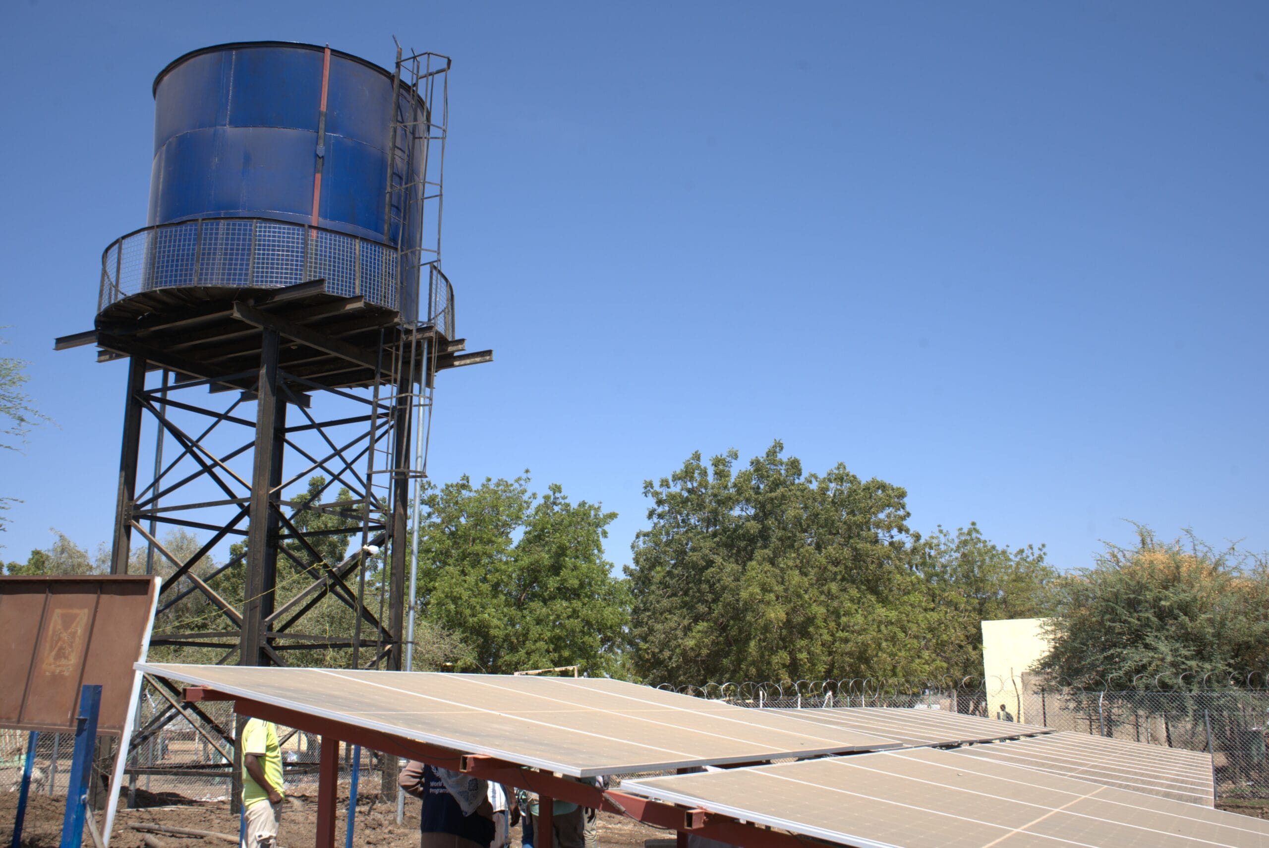 A blue elevated water tank stands on a metal frame beside several solar panels angled towards the sun, with trees and a clear blue sky in the background.