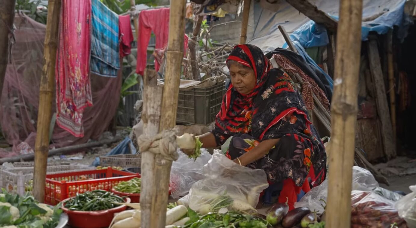 A woman in a colourful headscarf sits at an outdoor market stall, sorting and selling fresh vegetables such as aubergine, chilli peppers, and gourds, surrounded by makeshift wooden poles and fabric coverings.
