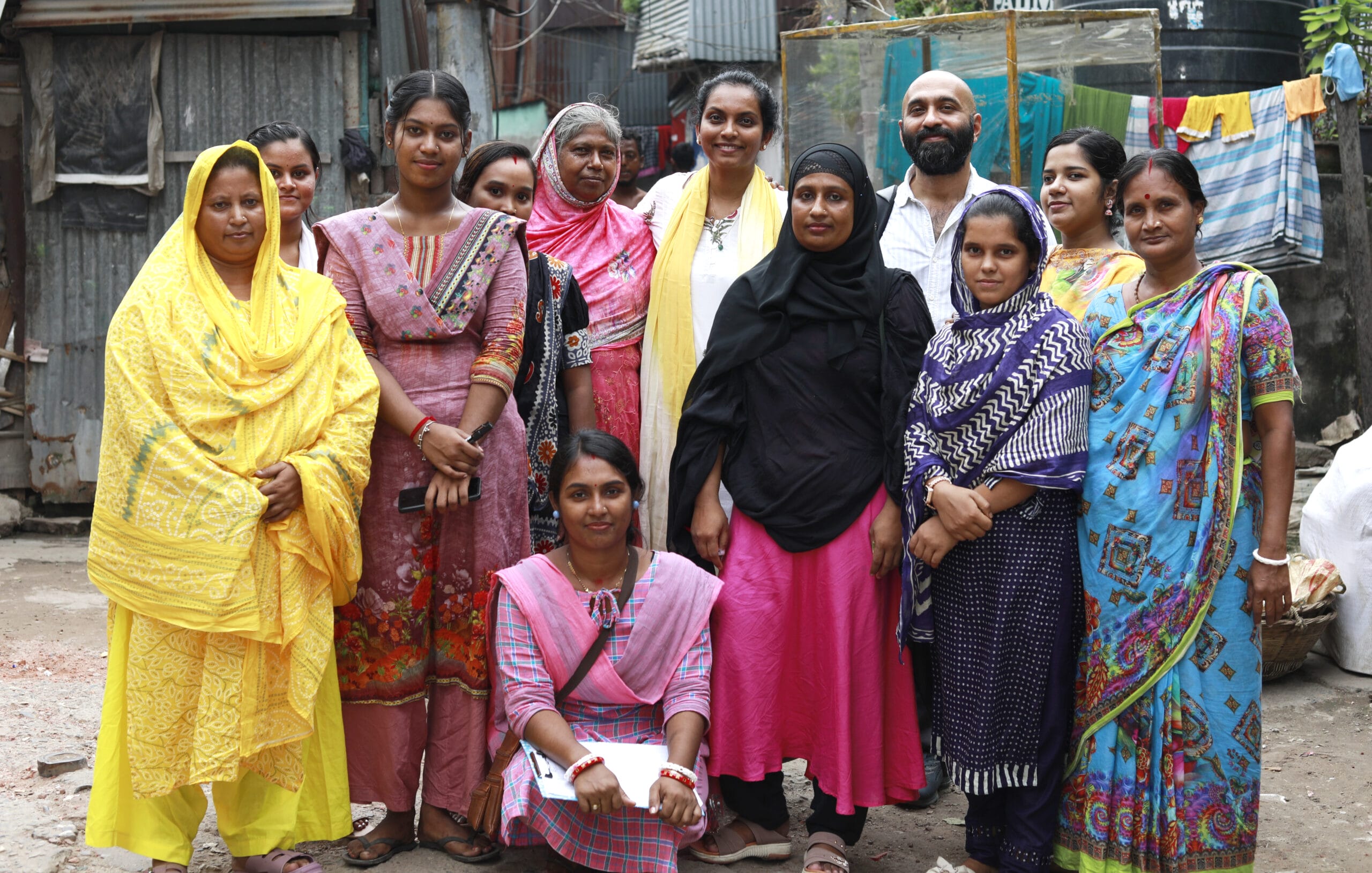 A group of thirteen people, mostly women in colourful traditional clothing, pose together outdoors in an urban area with corrugated metal and laundry hanging in the background.