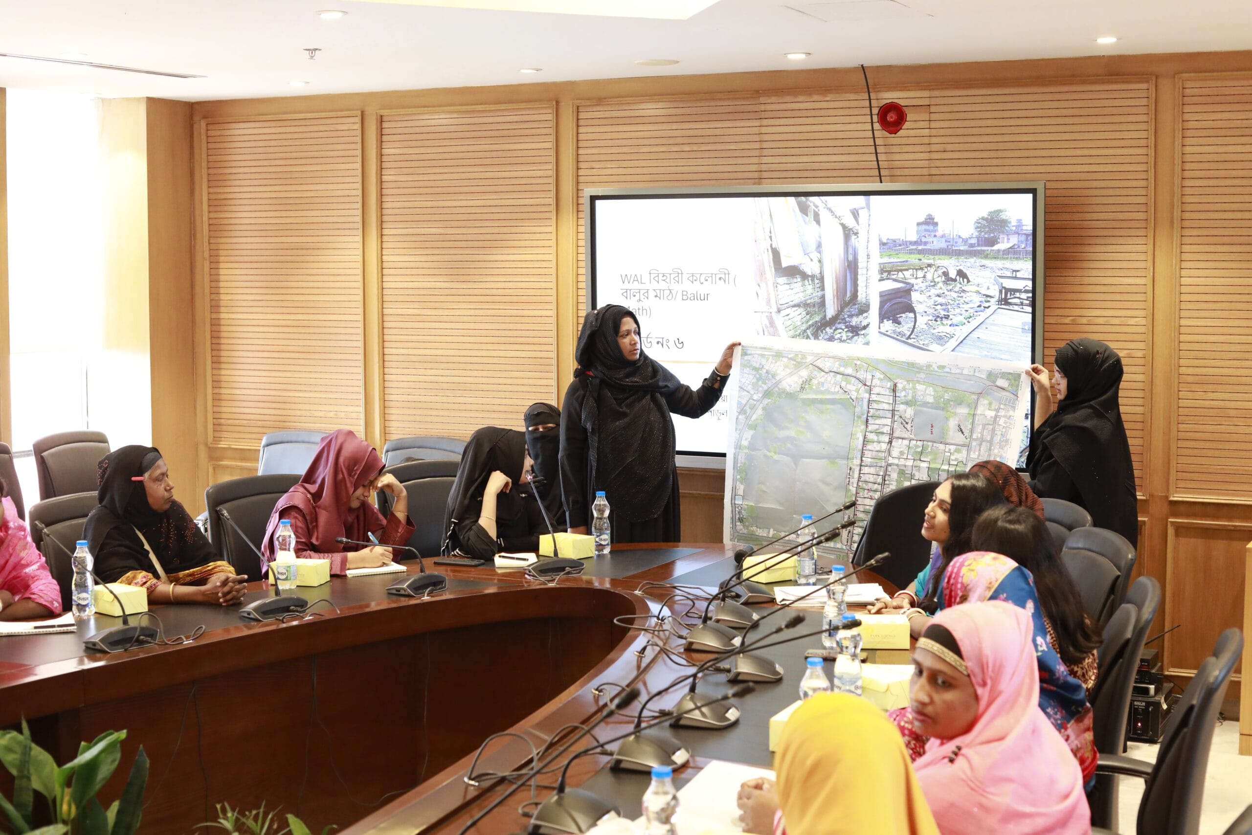 A group of women in hijabs sit around a conference table. Two women stand at the front, holding and discussing a large map. A screen behind them displays images and text.