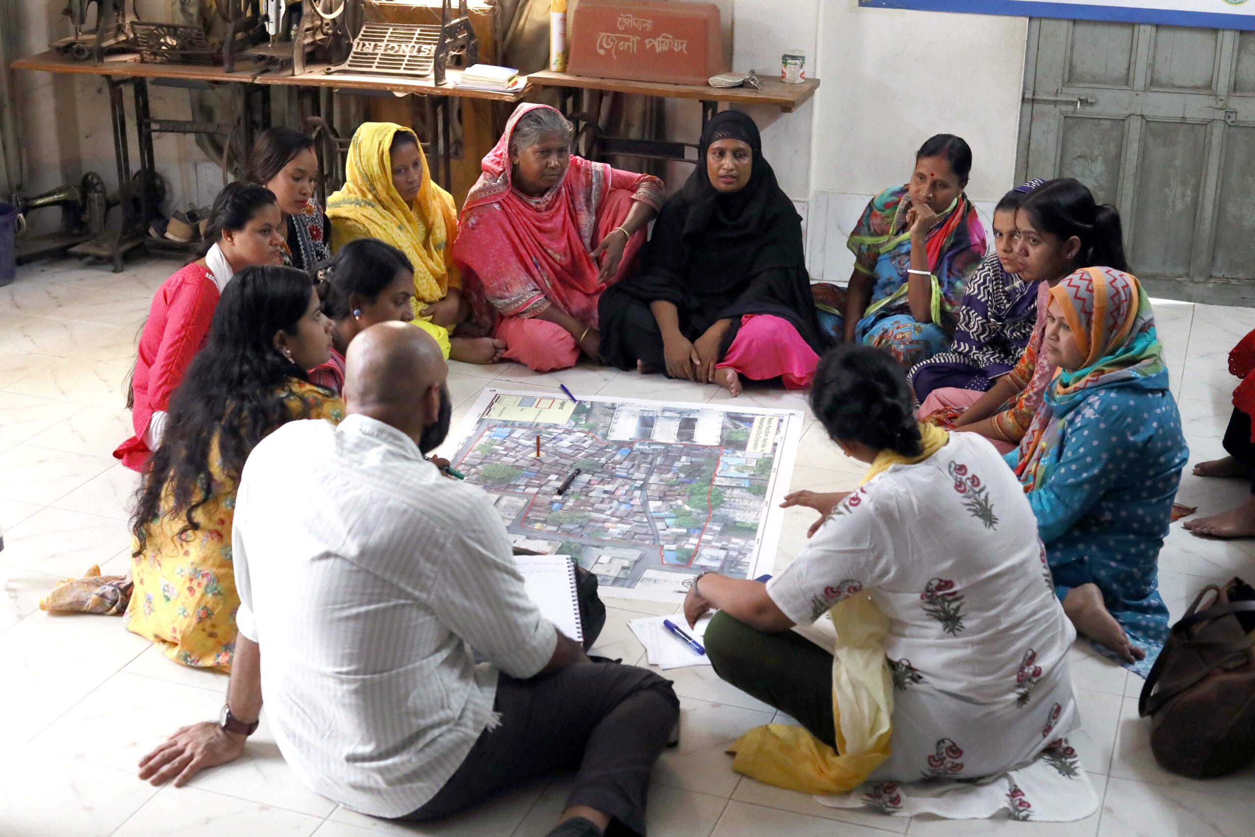 A group of women and a man sit in a circle on the floor, discussing and pointing at a large map or plan spread out in the centre, in a brightly lit indoor setting.
