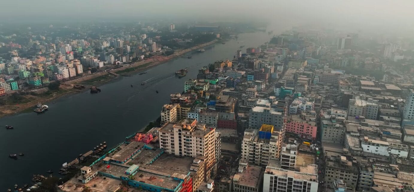 Aerial view of a densely populated city with colourful buildings along a wide river, under a hazy sky. Some boats are visible on the water, and the city extends into the distance.
