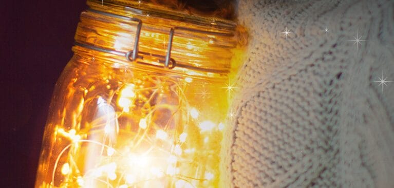A close-up of a person in a cream knitted jumper holding a glass jar filled with glowing fairy lights, with soft, warm light illuminating the scene.
