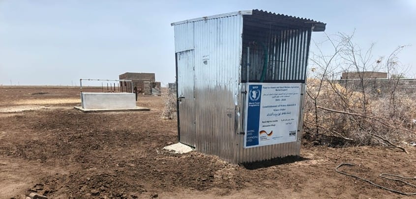 A small metal shed with a large sign stands on bare, dry soil in a rural area, with a concrete well and sparse bushes nearby under a hazy sky.