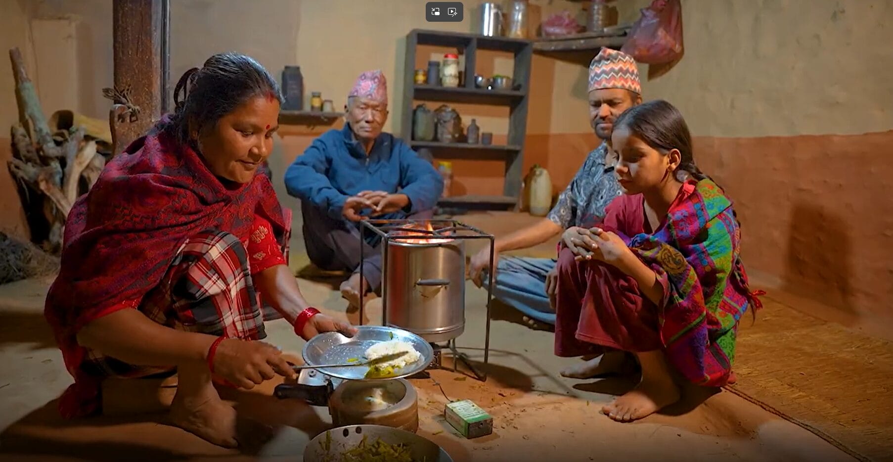 A woman cooks food on a cooker while two men and a girl sit nearby on the floor, inside a modest, warmly lit kitchen with shelves holding jars and utensils in the background.