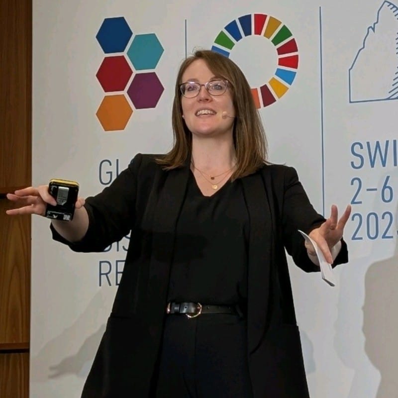 A woman wearing glasses and a black outfit is speaking on stage with a headset microphone. Behind her are colourful logos and event details, including “SWITZ 2-6 2023.” She gestures with her hands whilst presenting.