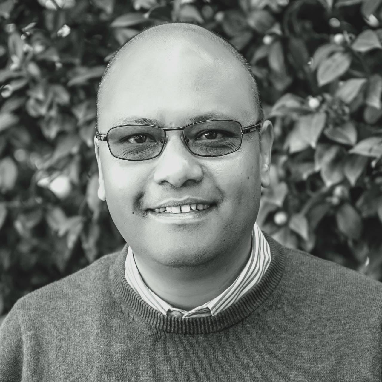 A person with short hair and glasses smiles at the camera, wearing a jumper over a collared shirt. The background features leafy foliage. The photo is in black and white.
