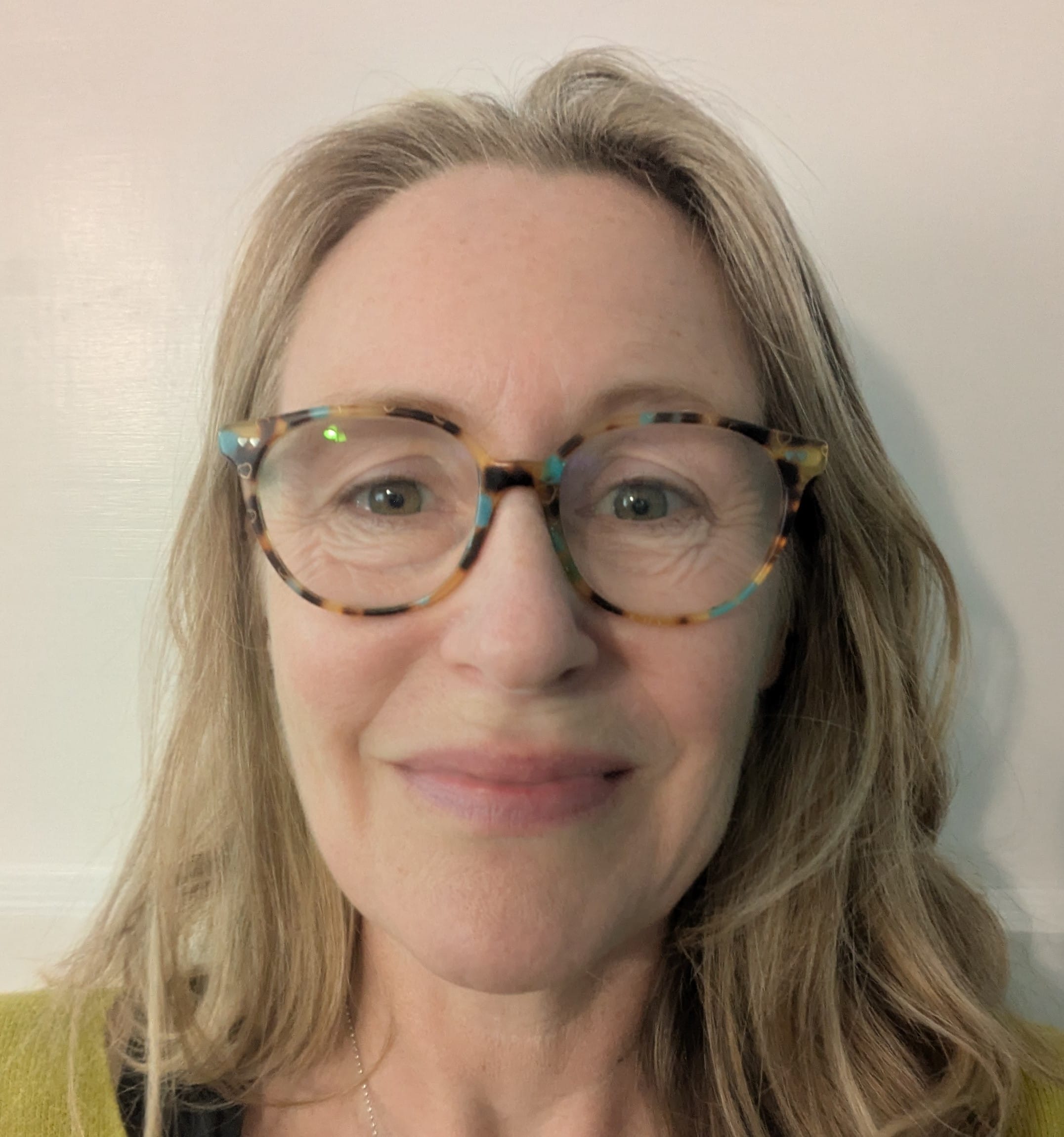 A woman with long, light brown hair and tortoiseshell glasses smiles softly at the camera against a plain, light-coloured background.
