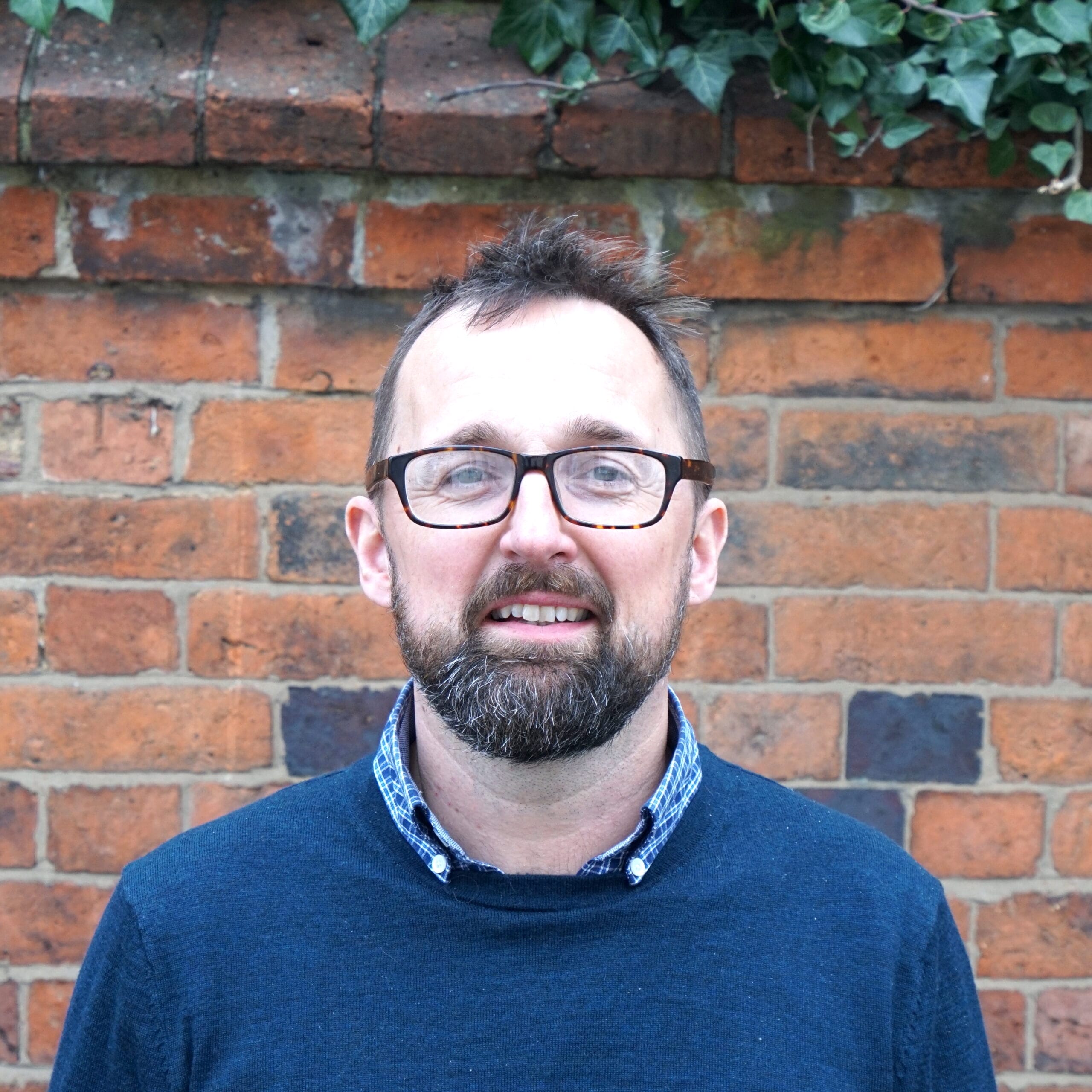 A man with glasses and a beard is standing in front of a brick wall, wearing a dark blue jumper over a patterned shirt. Green ivy hangs over the top of the wall.