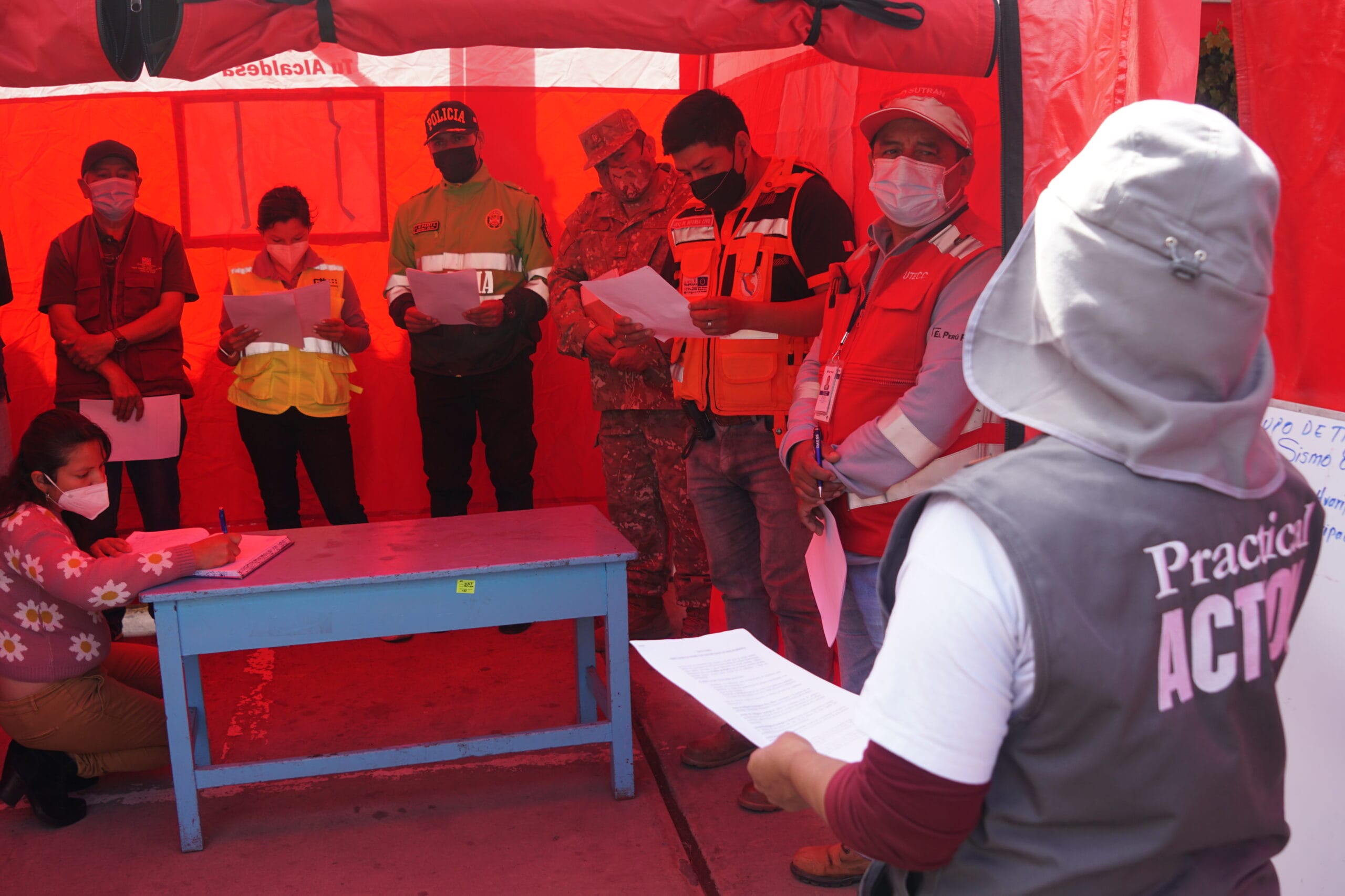 A group of people in safety vests and uniforms, some wearing masks, stand inside a red marquee holding papers, appearing to participate in an emergency drill or briefing. One person writes at a blue desk.