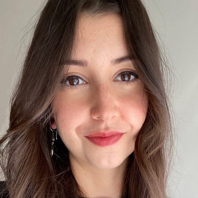 A young woman with long brown hair, brown eyes, and light skin smiles softly at the camera. She wears subtle make-up and dangly earrings, with a plain light background behind her.
