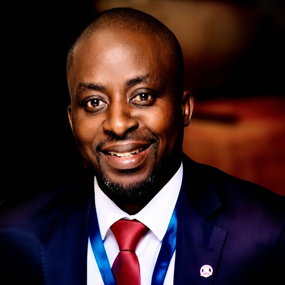 A smiling man in a dark suit, white shirt, and red tie, wearing a blue lanyard and a lapel pin, poses for a professional portrait against a dark, blurred background.